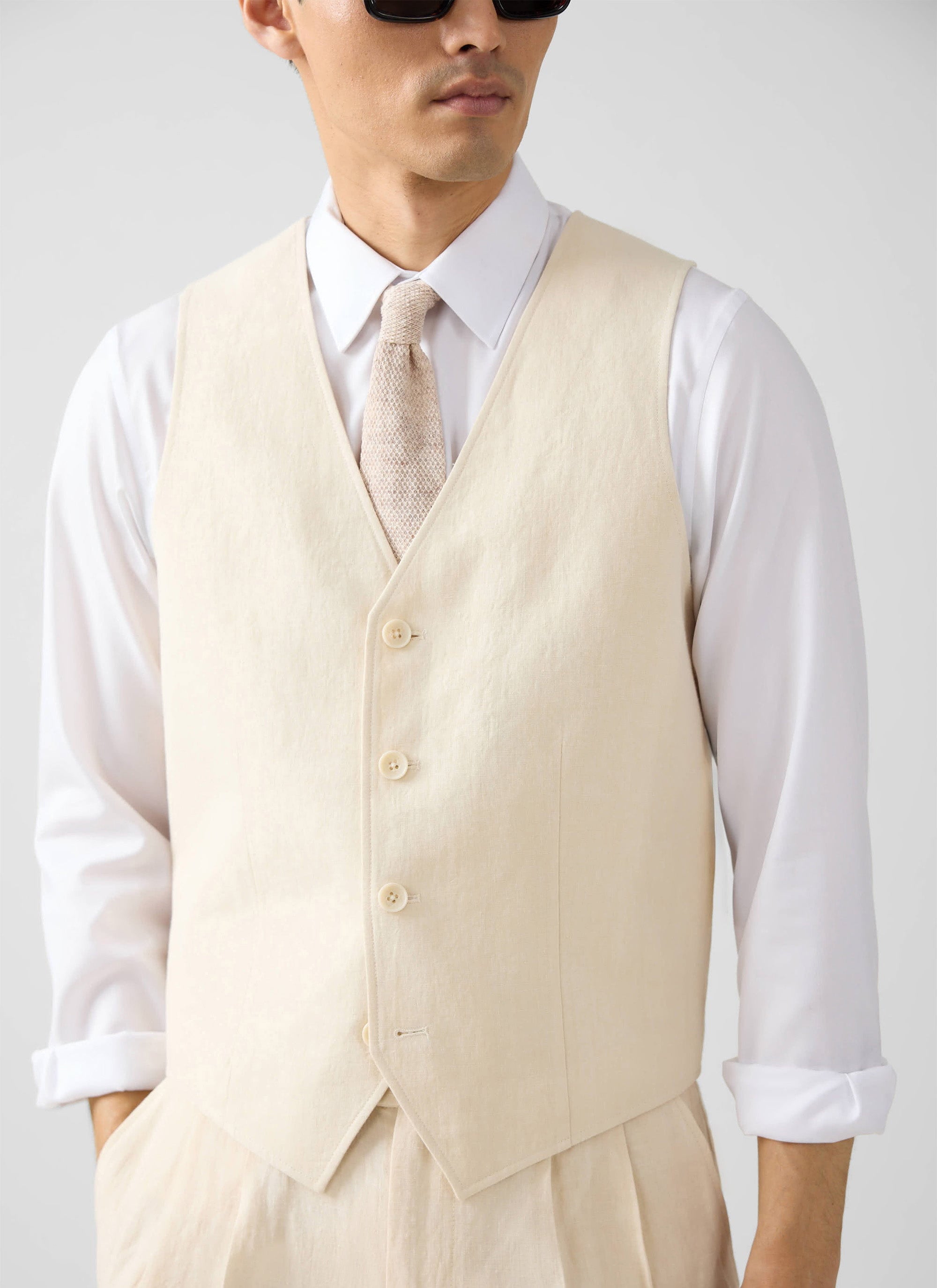 A model wears the Percival cream linen waistcoat, styled with a white shirt, textured tie, and matching linen trousers.