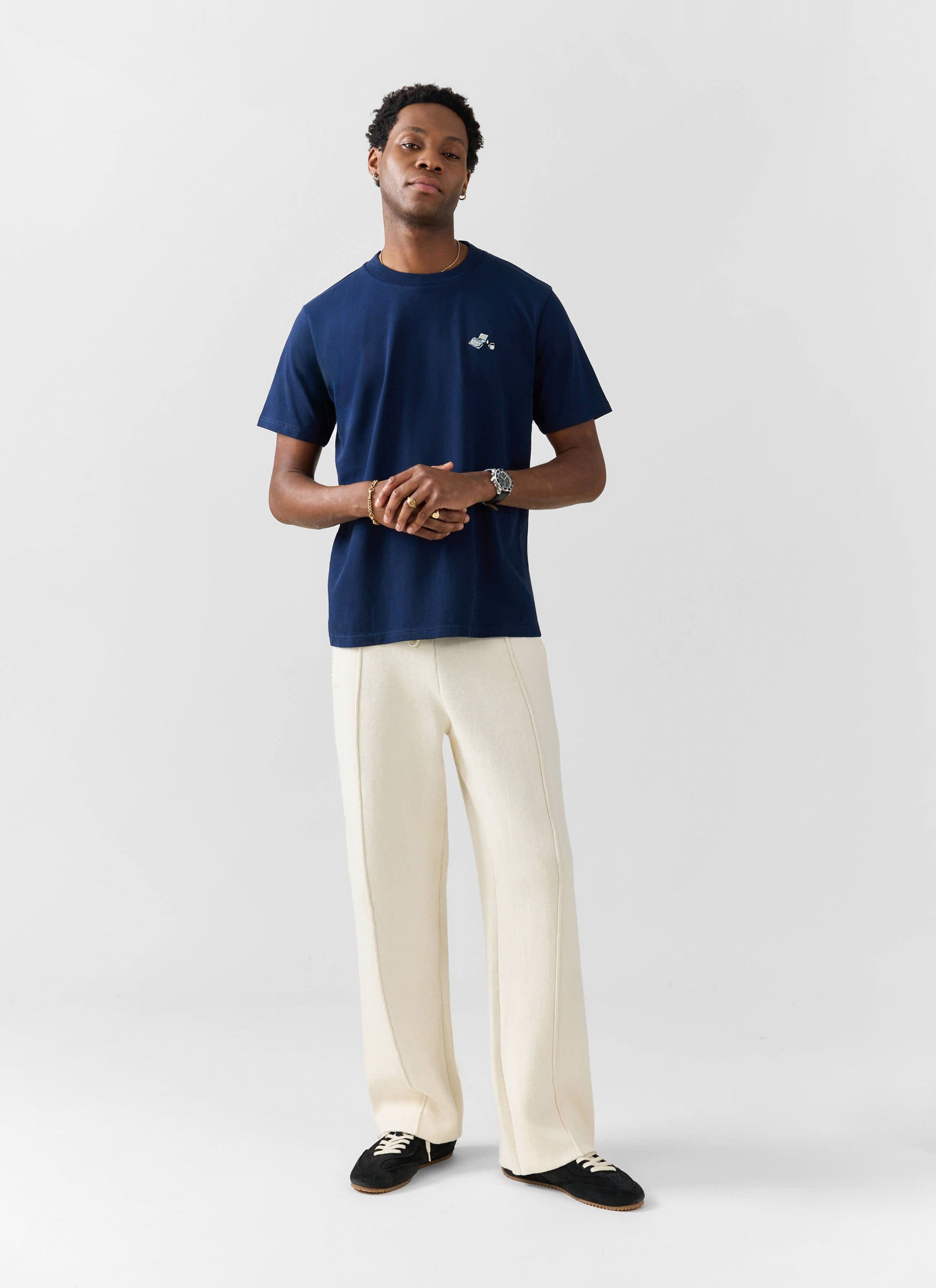 A model wears the Percival Typewriter Embroidered T Shirt in navy with wide-leg cream trousers and black trainers.