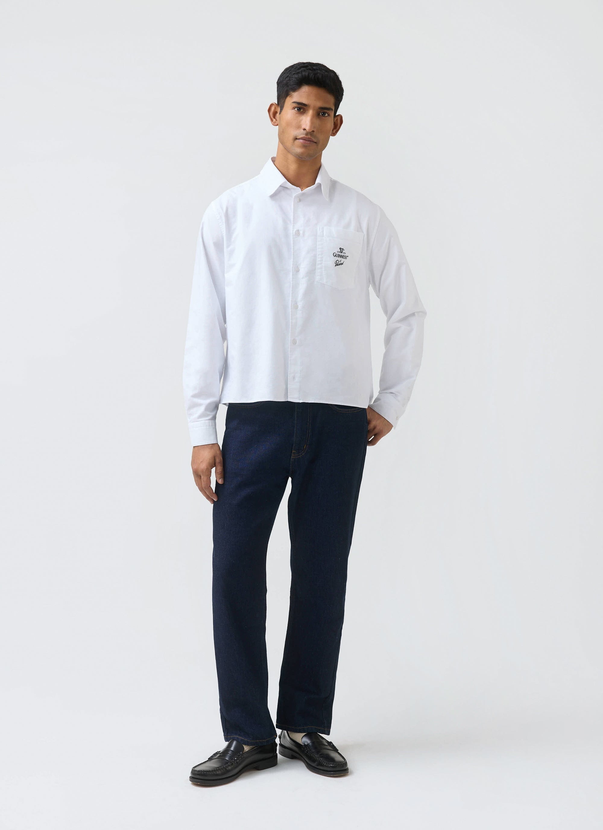A man models the white Guinness x Percival Button Down Boxy Shirt, styled with dark jeans against a plain background.