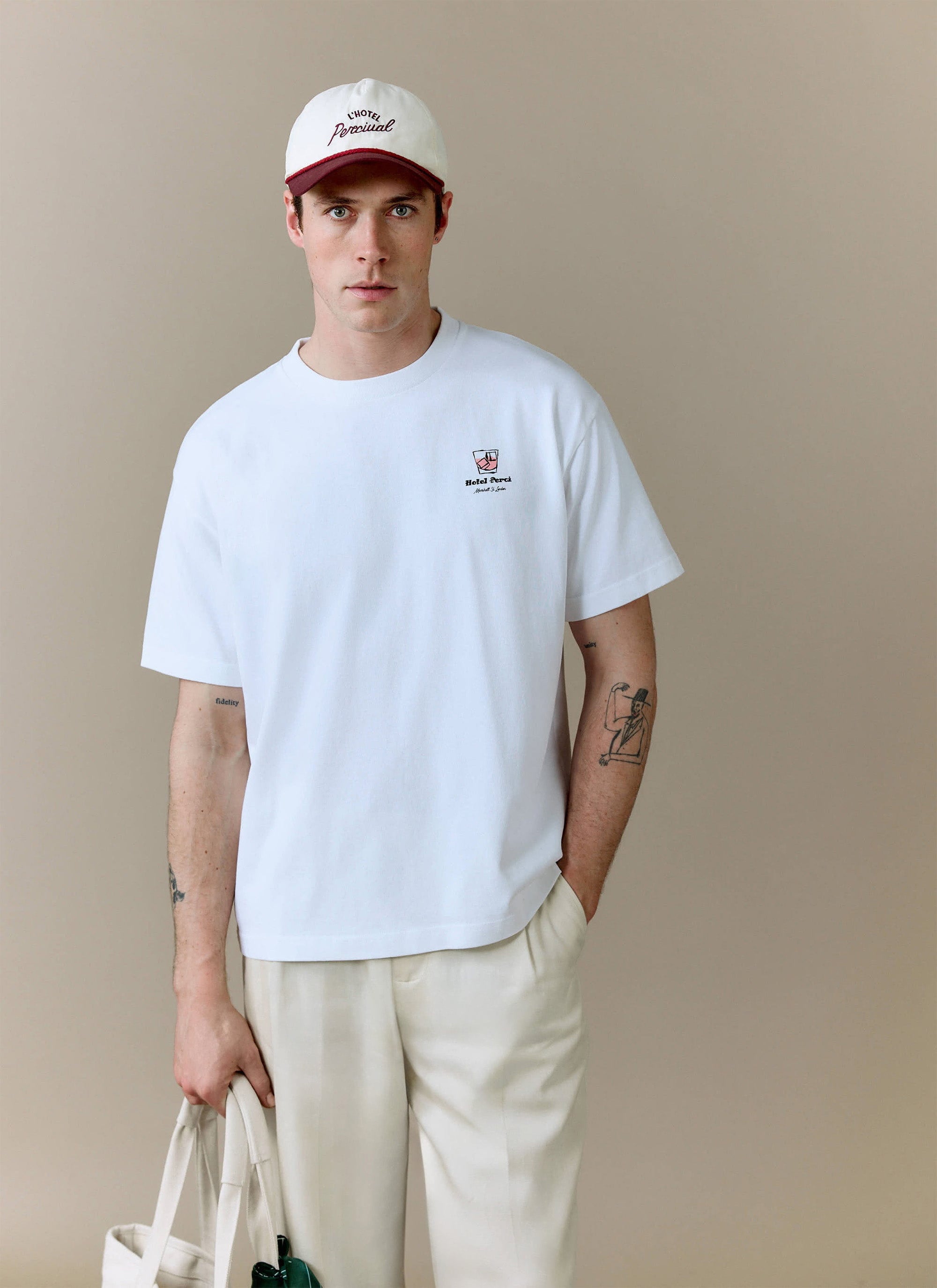 A model wears the white Hotel Bistro Oversized T-Shirt from Percival, styled with light trousers and a branded cap.