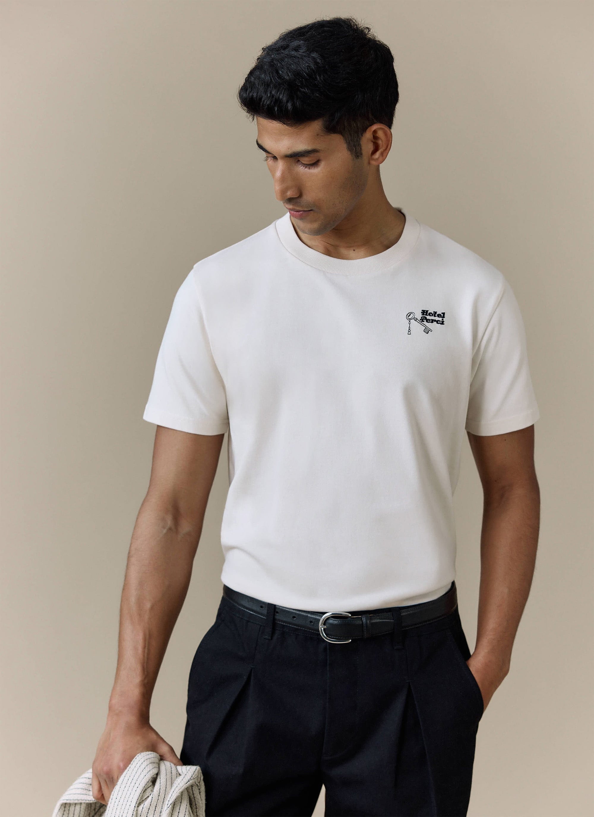 A model wears the Percival Hotel T-Shirt in ecru organic cotton, styled with smart black trousers.