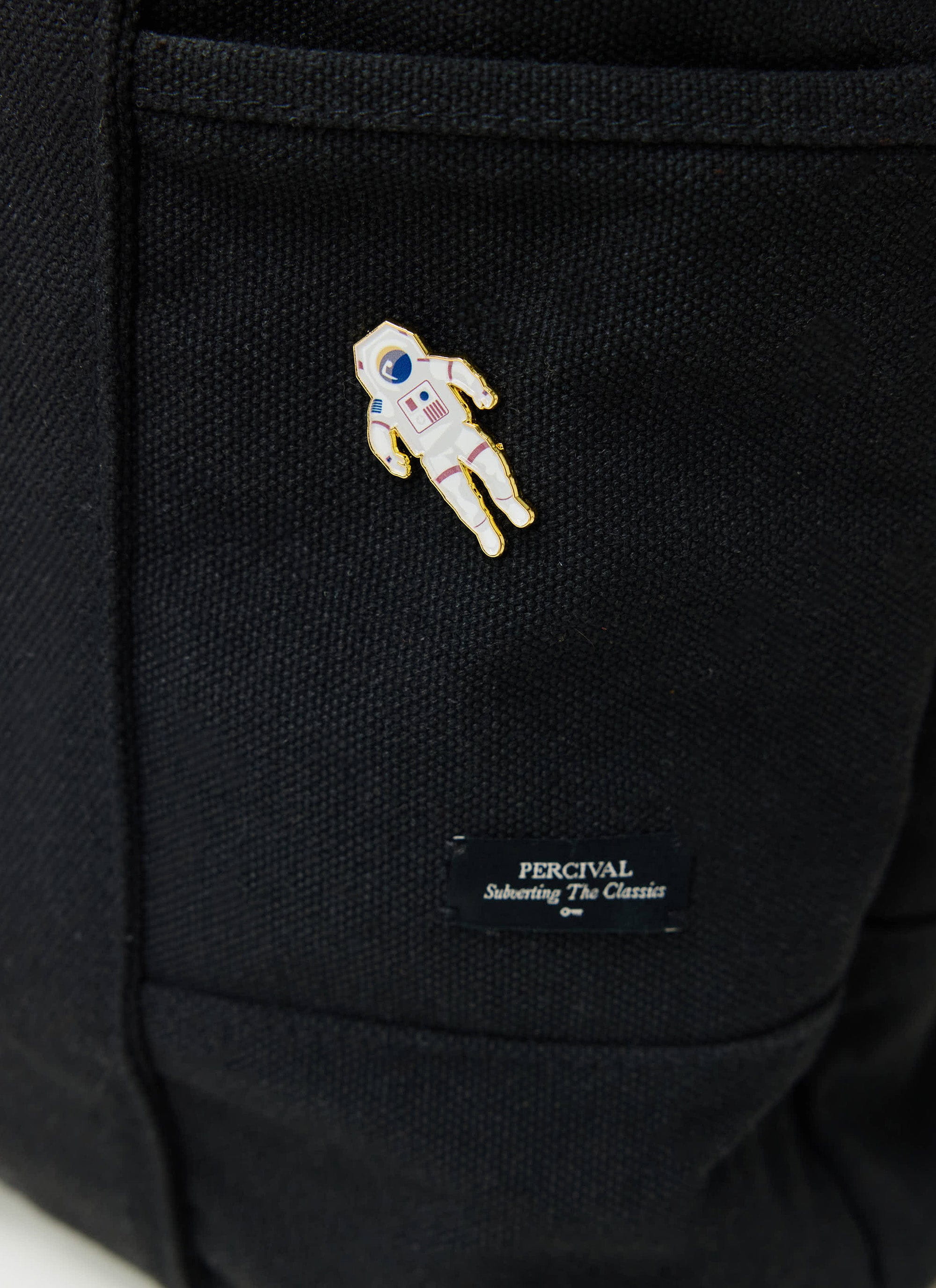 A close-up of the Percival Spaceman Pin Badge pinned onto a black jacket, just above the Percival brand tag.