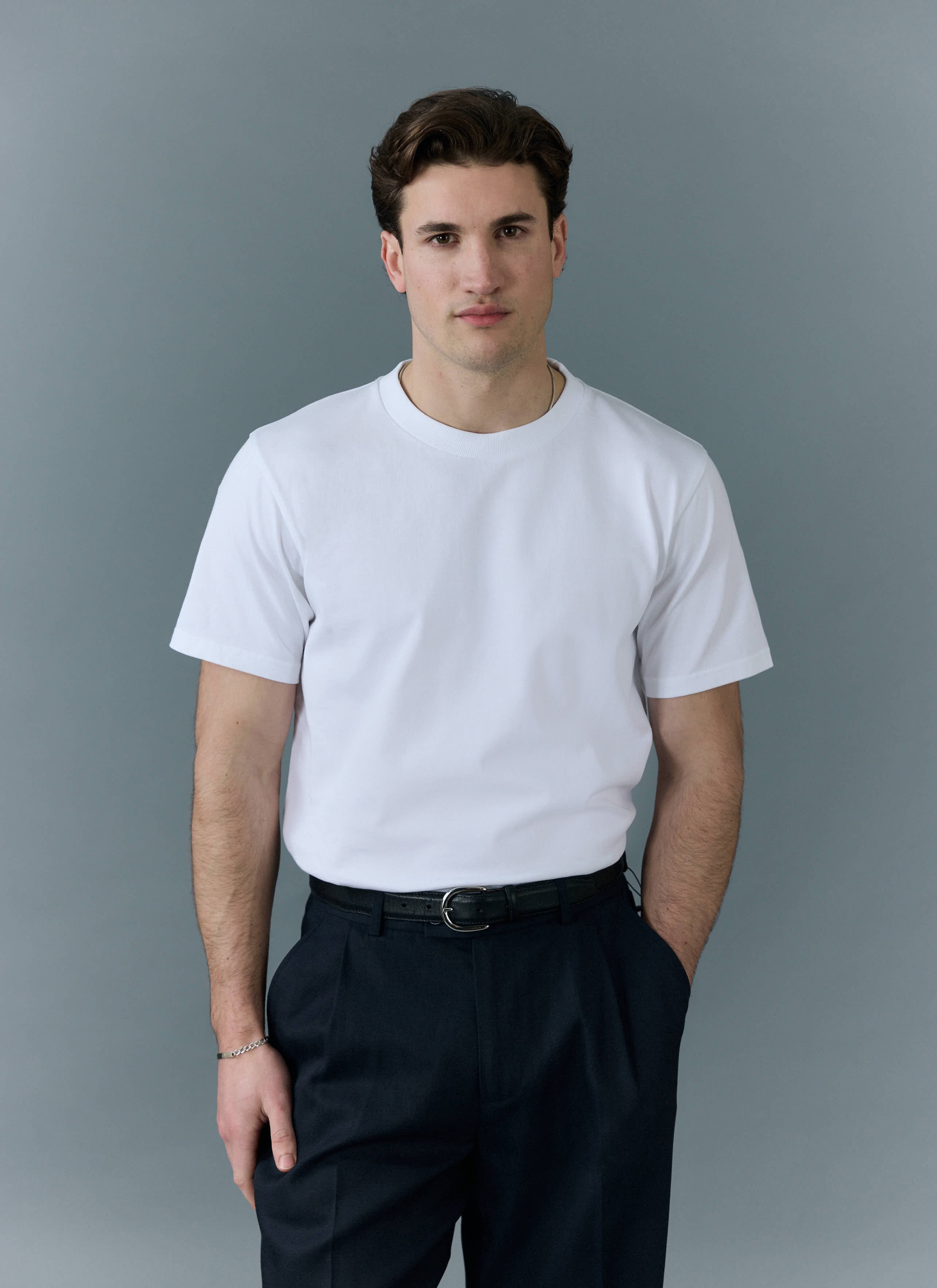 A model wears the Percival men's plain white organic cotton t-shirt with dark trousers against a grey background.