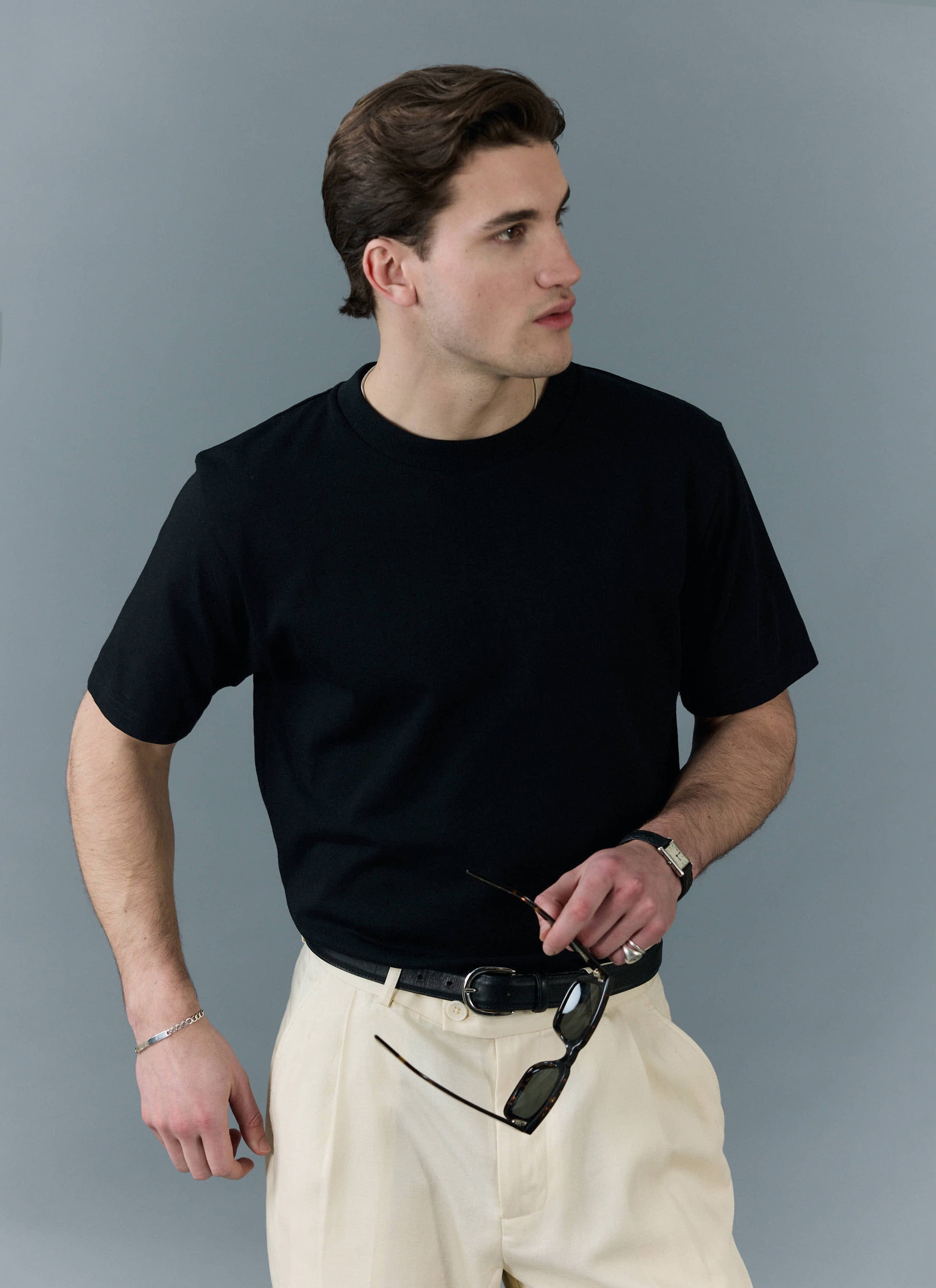A man models the Percival black organic cotton t-shirt, styled with cream trousers and sunglasses.