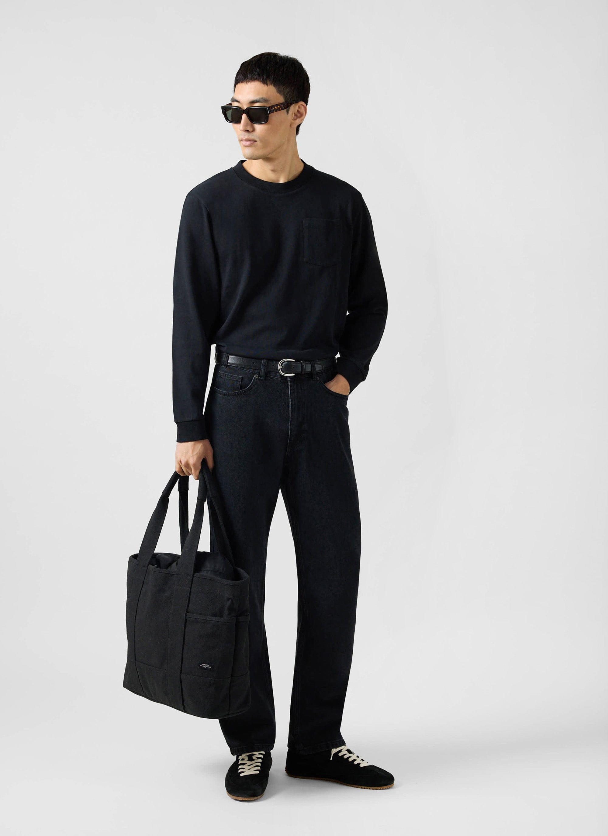 A model wears the Percival black Organic Cotton Long Sleeve Pocket T Shirt, with black jeans, sunglasses and a tote bag.