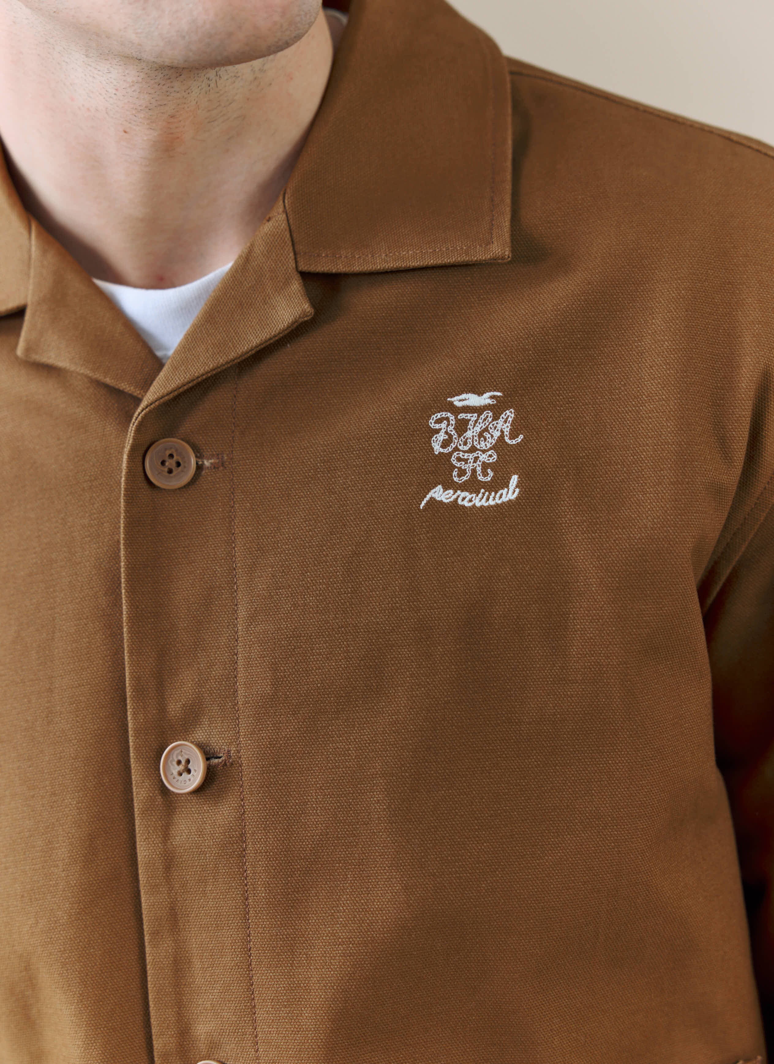 A close-up of the white embroidered logo on the chest of the Percival x Brighton & Hove Albion FC Canvas Workshirt in brown.