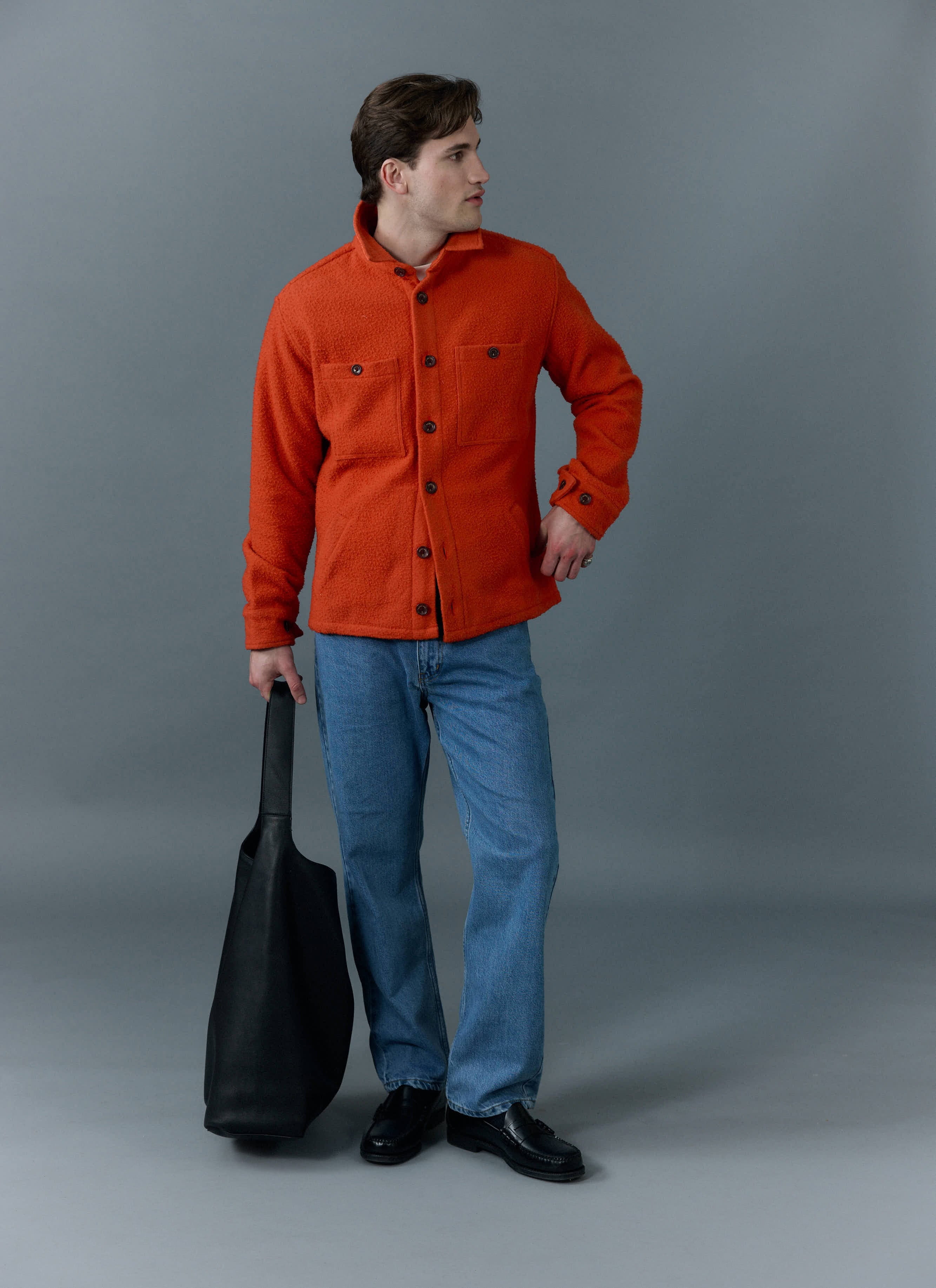 A model wears the Percival orange Casentino wool Blanket Overshirt with blue jeans and black loafers, holding a tote bag.