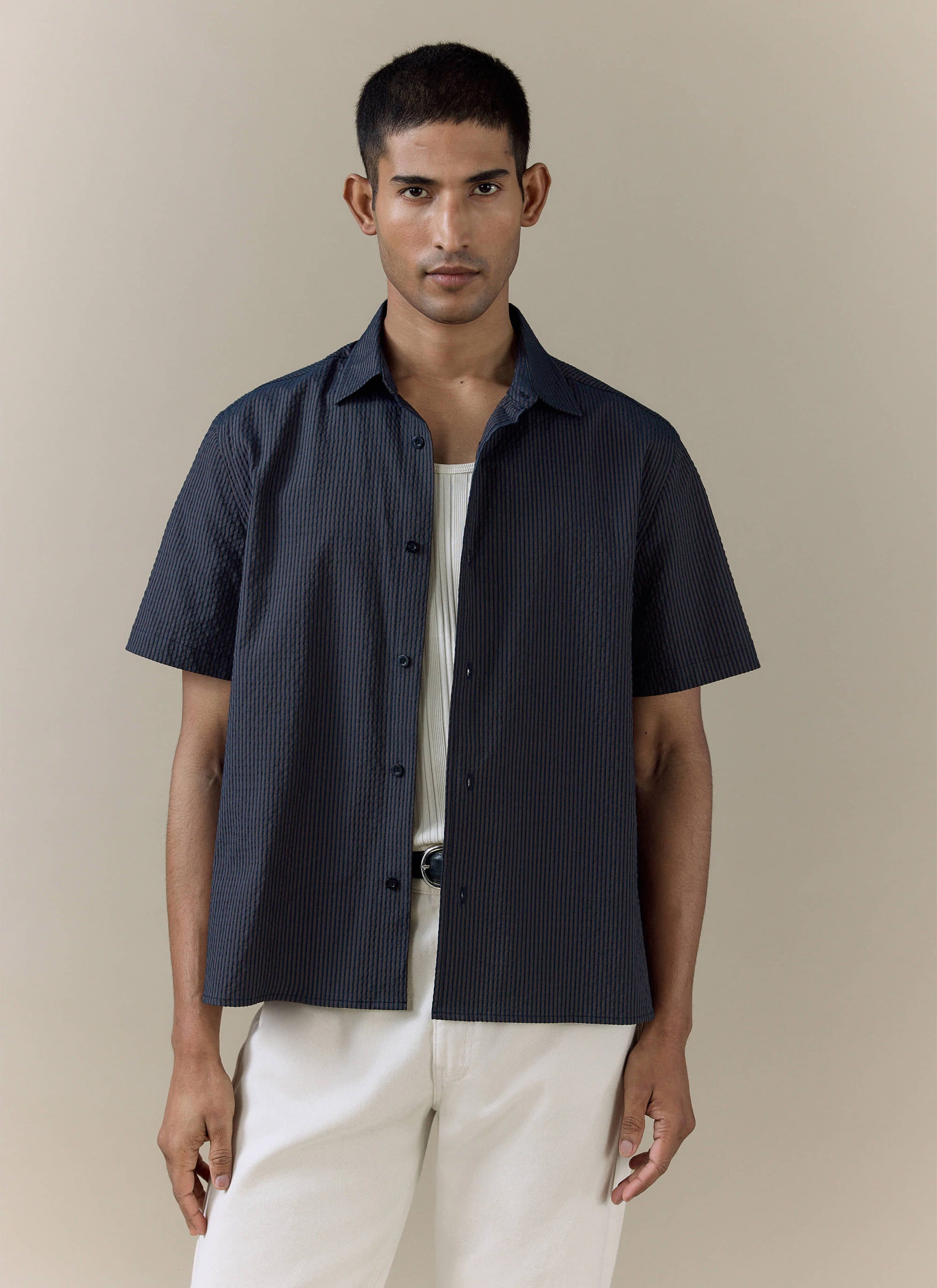 Percival Rowan Seersucker Boxy Shirt in navy stripe cotton, styled open over a white ribbed vest and cream trousers.