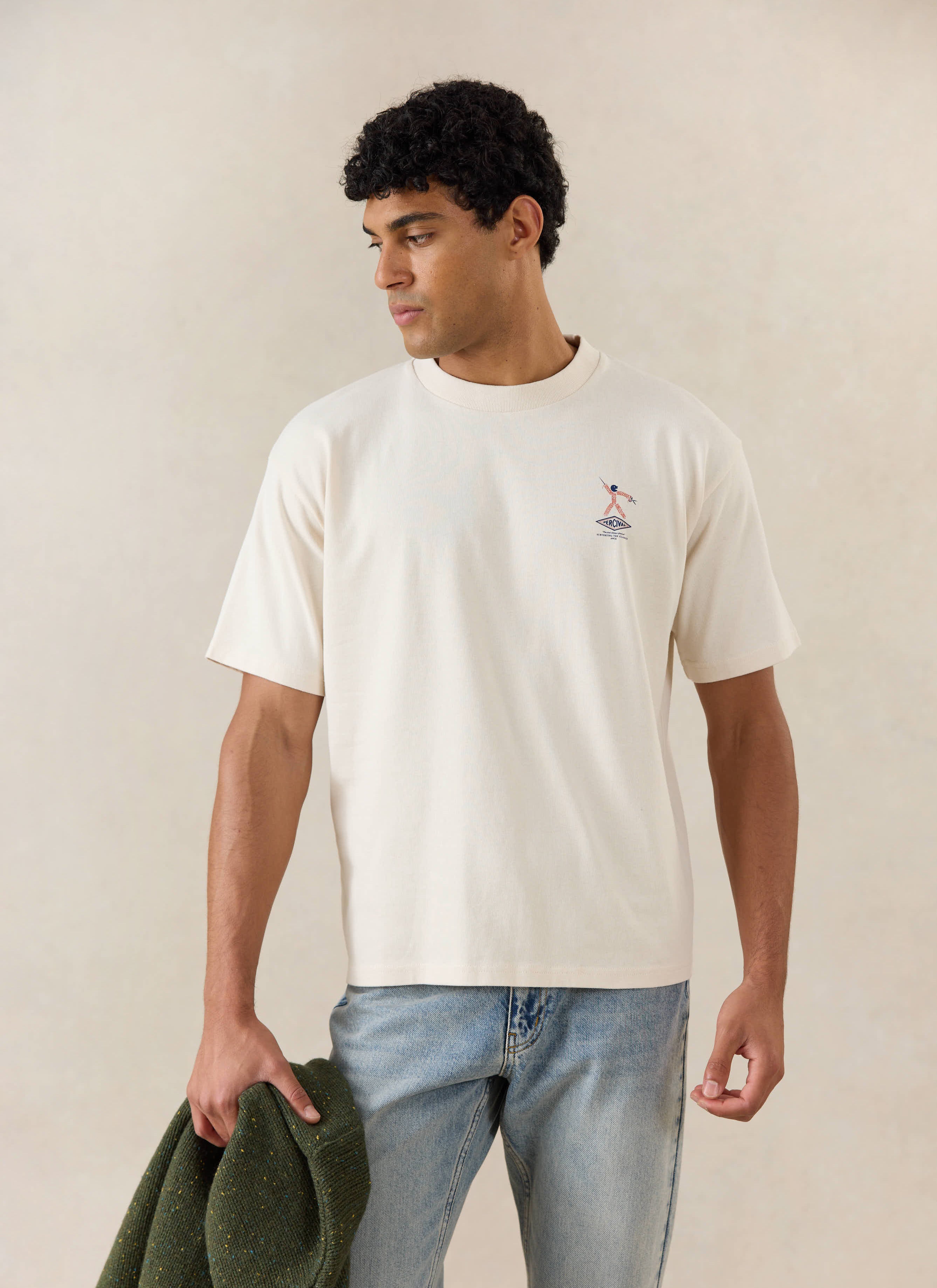 A model wears the cream Percival Kingston Boxy T-Shirt, styled with light blue jeans and holding a dark green knit.