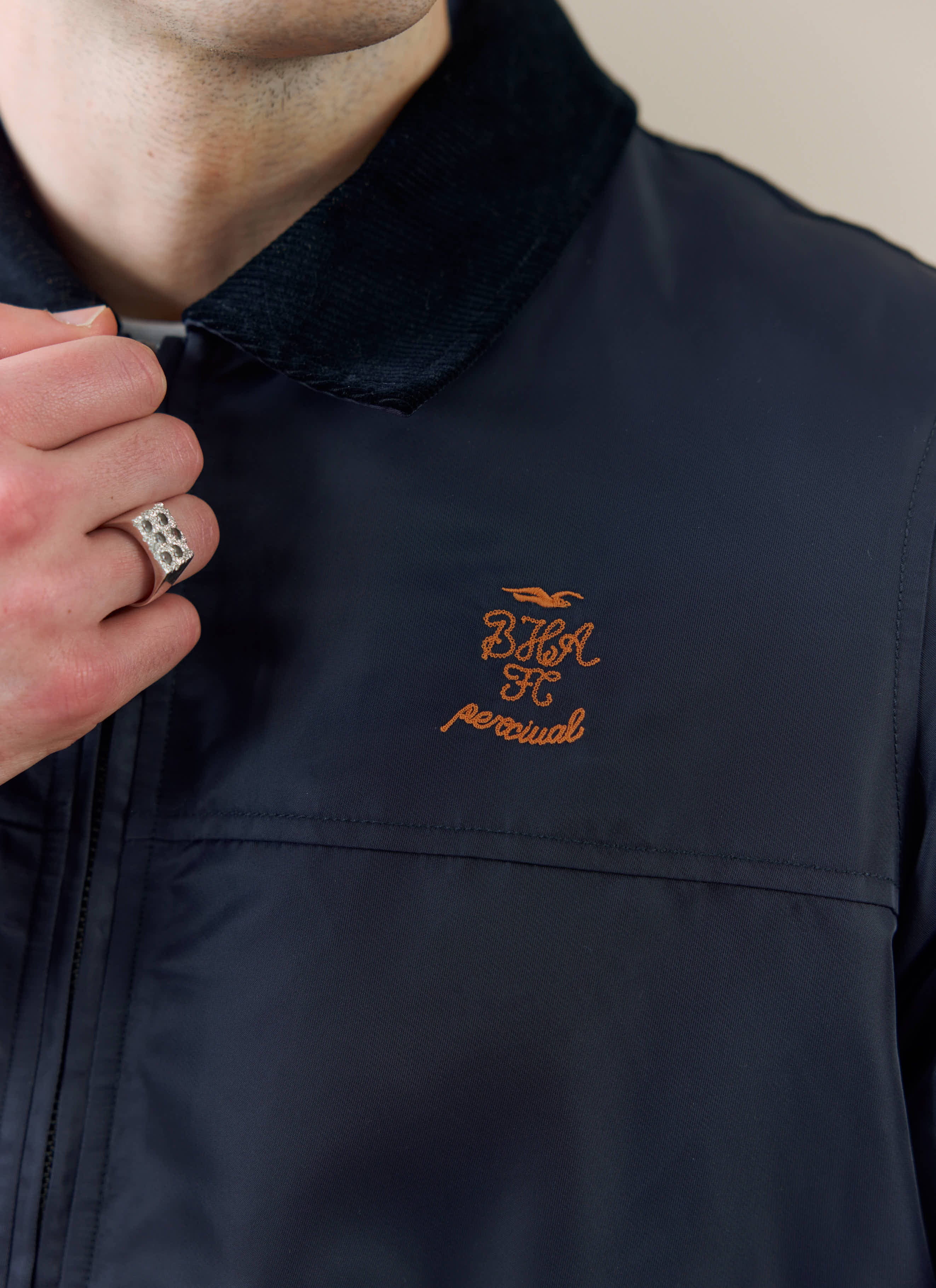 Close-up of the navy Percival x Brighton & Hove Albion coach jacket, showing the orange chest embroidery and cord collar.