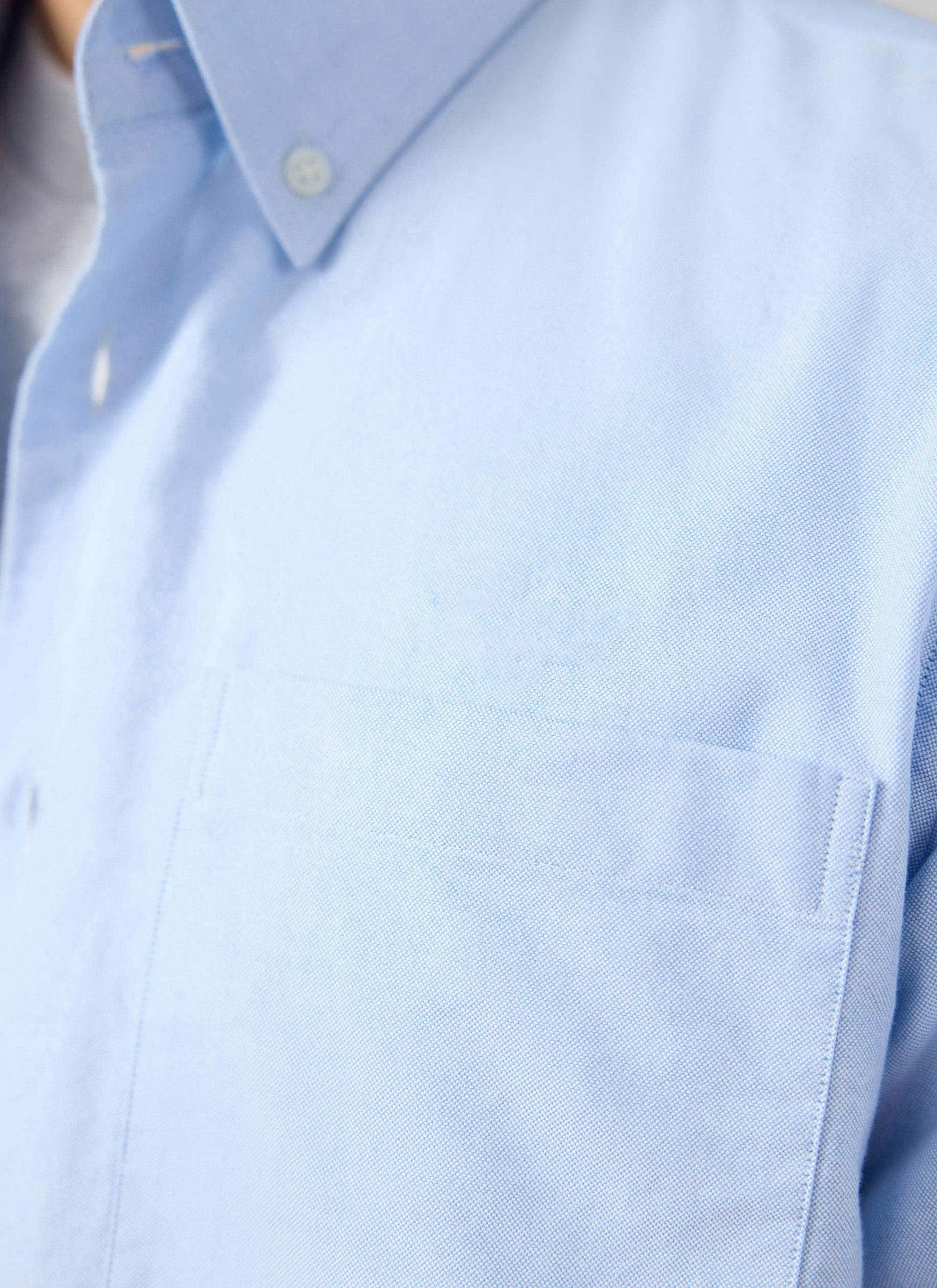A close-up detail shot of the Percival Oxford Regular Shirt in light blue, showing the chest pocket and button-down collar.