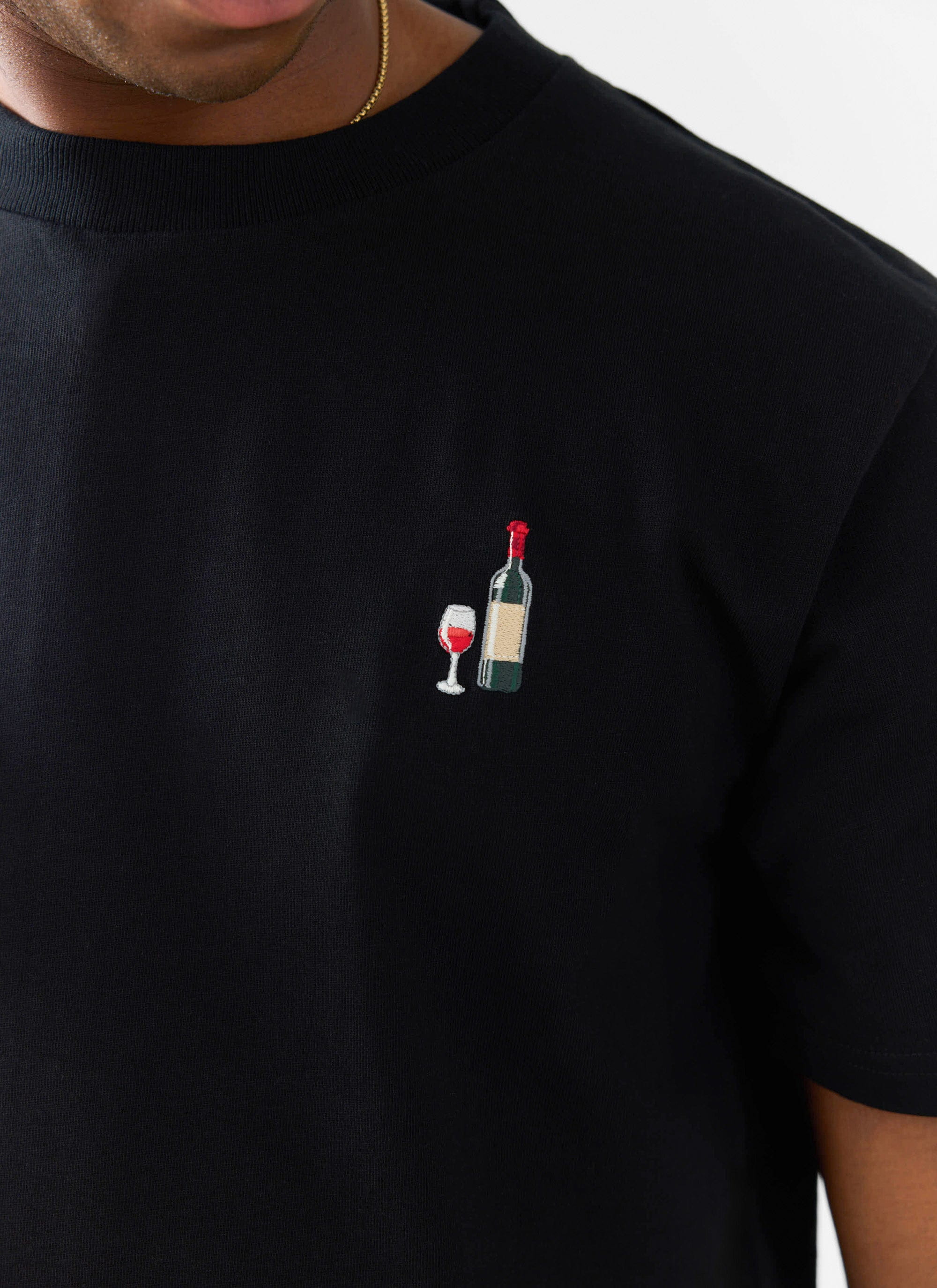 A close-up of the red wine bottle and glass embroidery on the chest of the black Percival t-shirt.