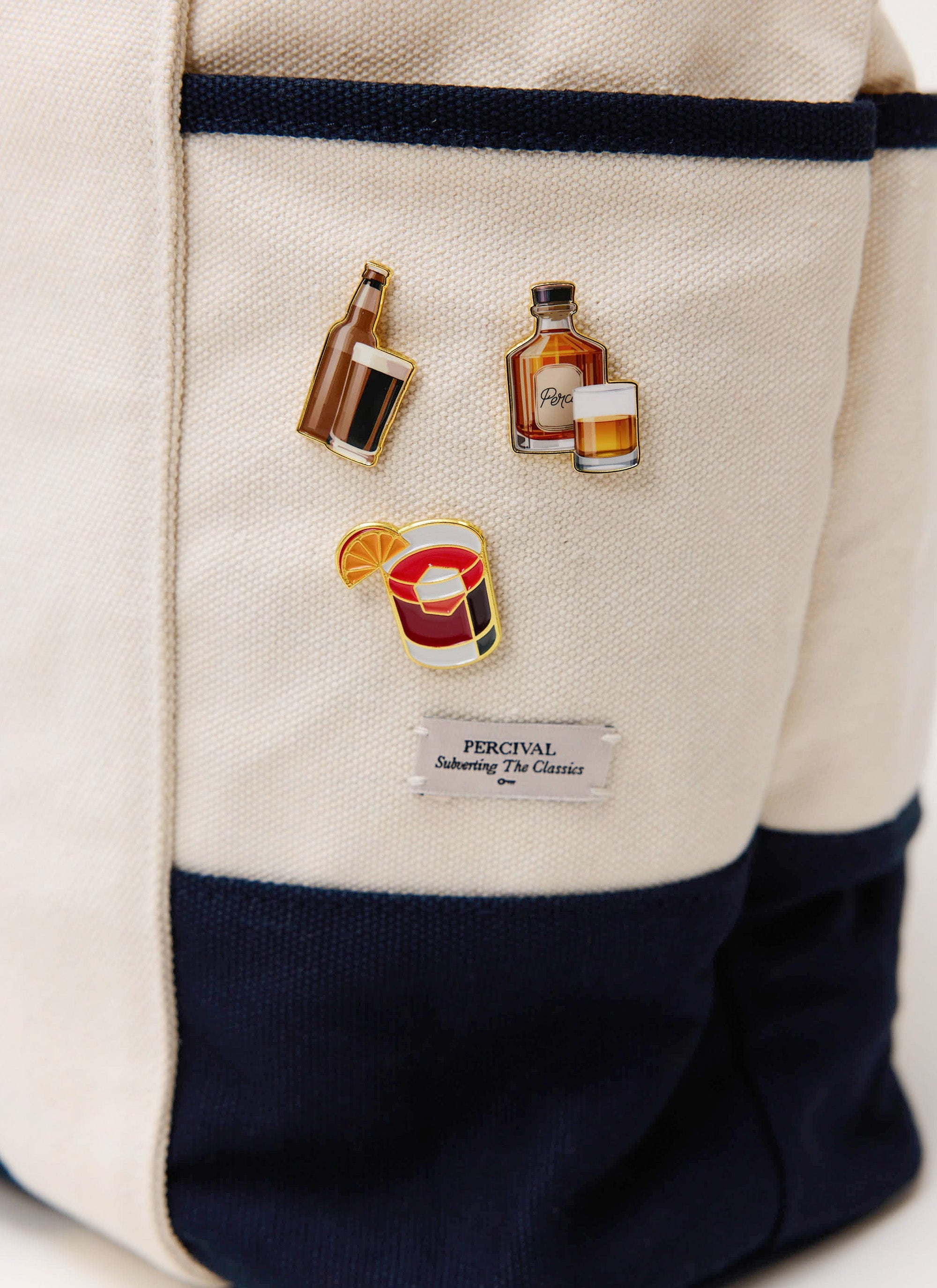 A close-up of a cream and navy canvas bag from Percival, adorned with the Stout, Whisky, and Negroni pin badges.
