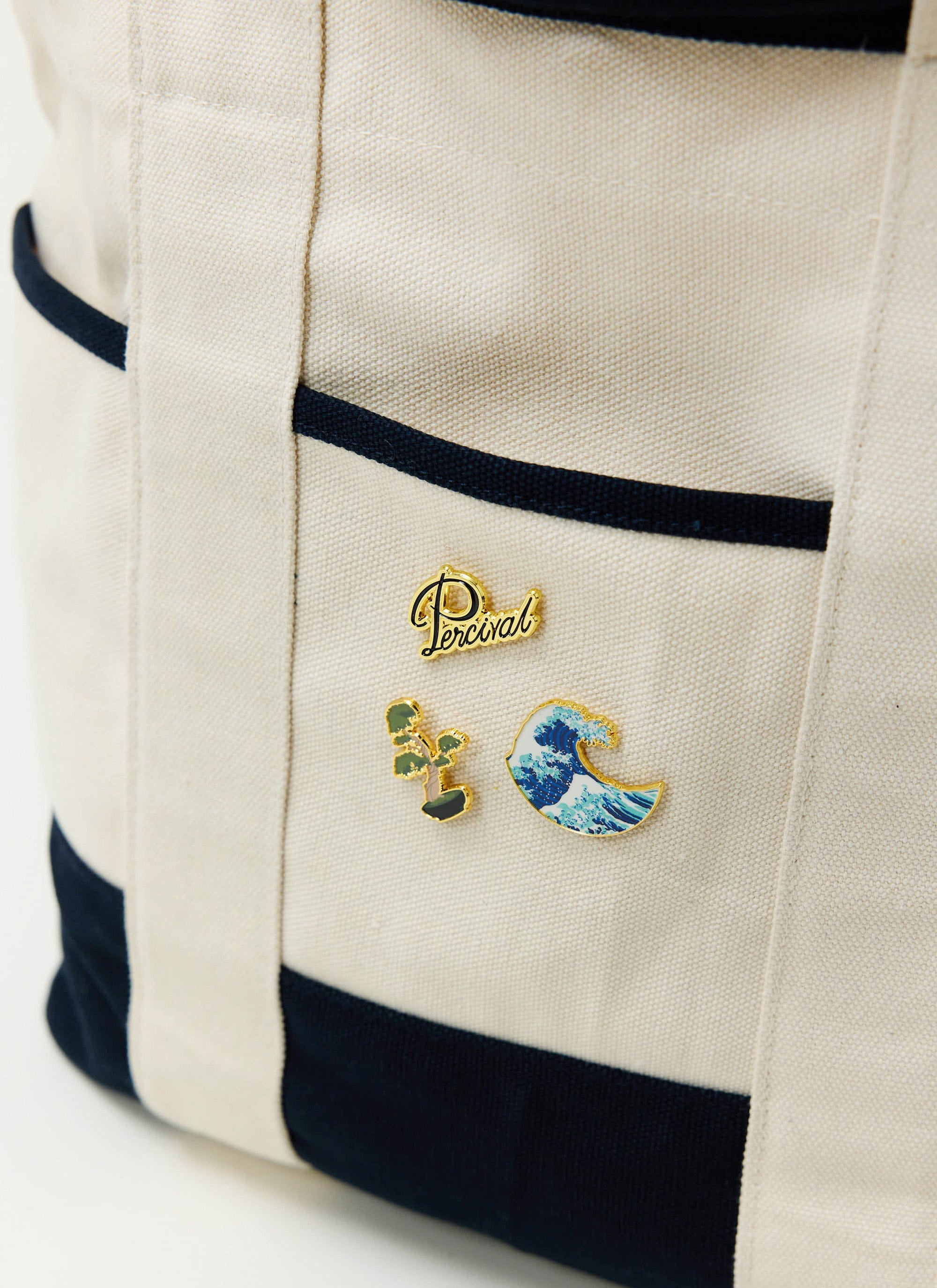 Three Percival pin badges, including a wave, a bonsai tree, and the Percival logo, are pinned to a cream canvas tote bag.
