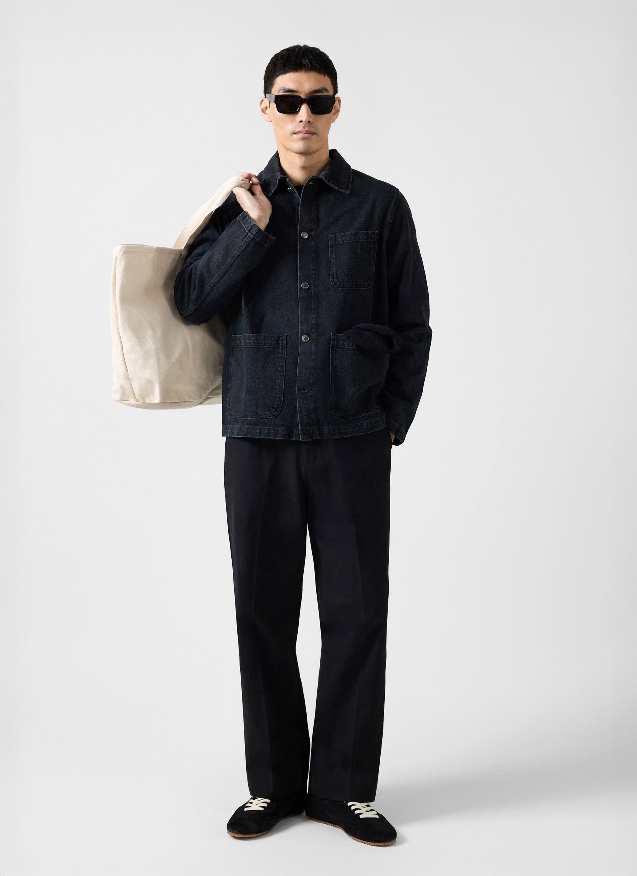 A model wears the Percival Gove Chore Jacket in black denim, styled with black trousers, sunglasses and a canvas tote bag.