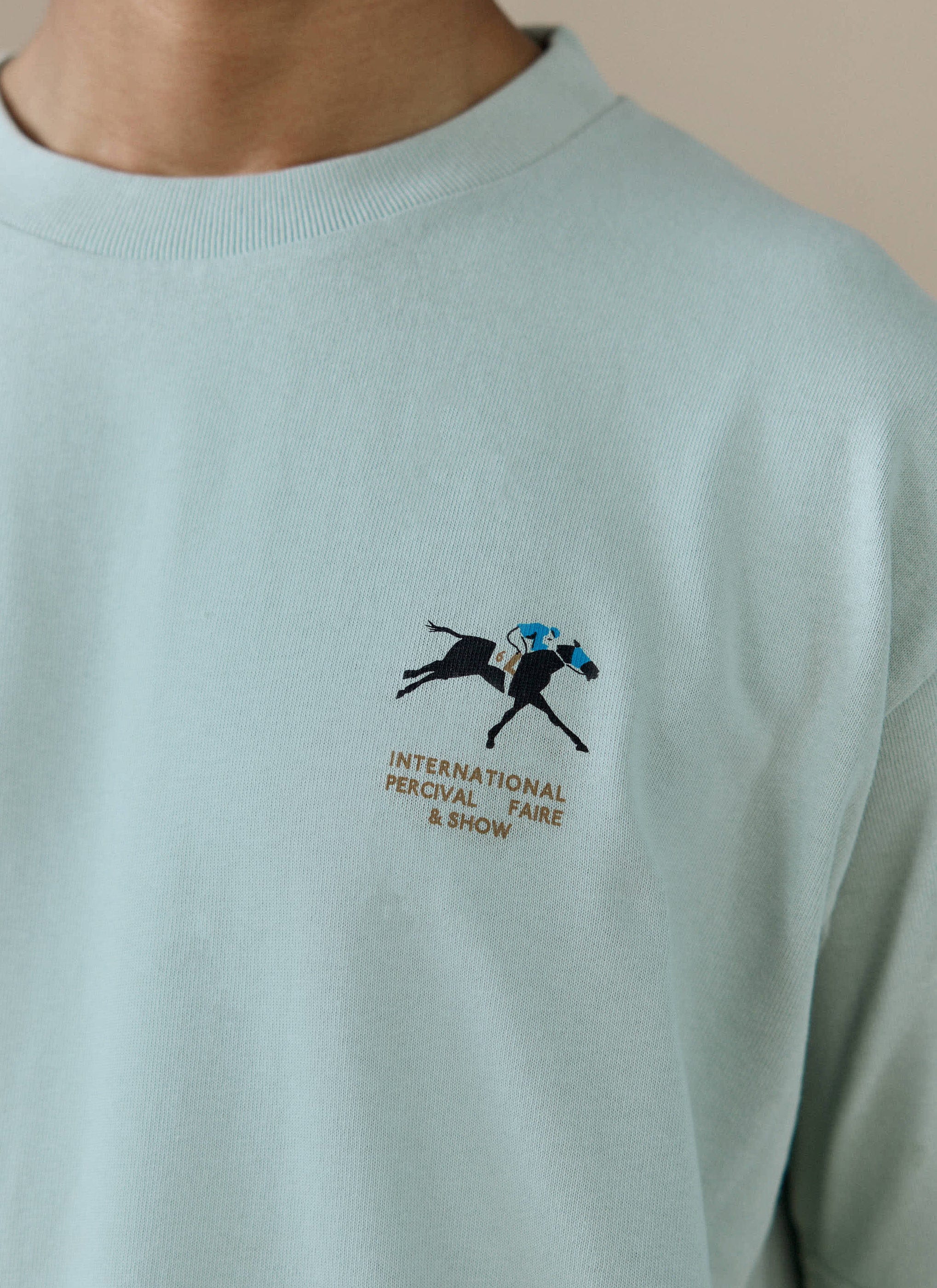 A close-up of the International Percival Faire & Show chest graphic on the sage green organic cotton oversized t-shirt.