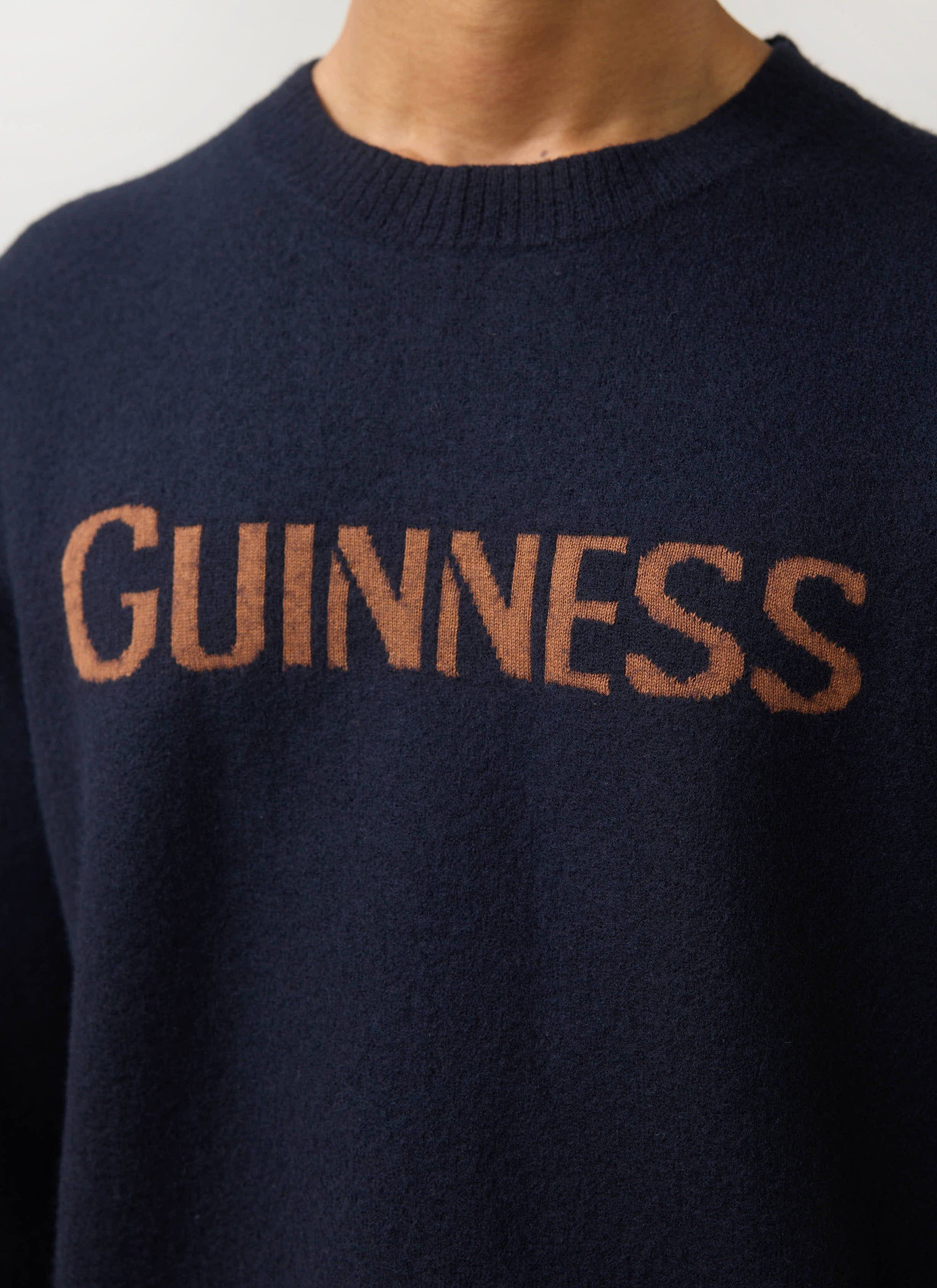 A close-up of the navy Guinness x Percival merino wool jumper, showing the chocolate jacquard 'GUINNESS' logo on the chest.