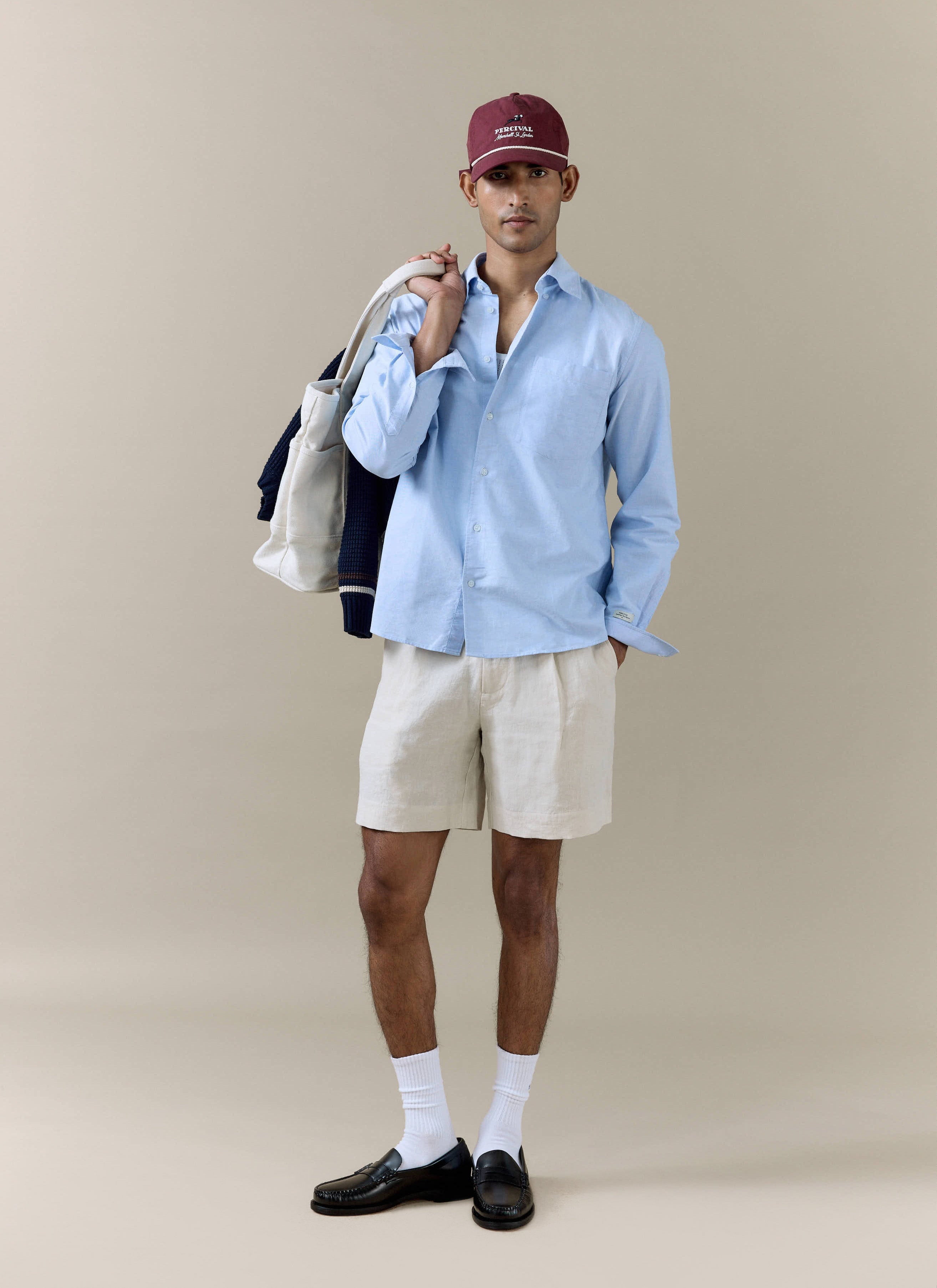 Cream Percival pleated linen shorts worn by a model with a light blue shirt, burgundy cap, and a white canvas tote bag.