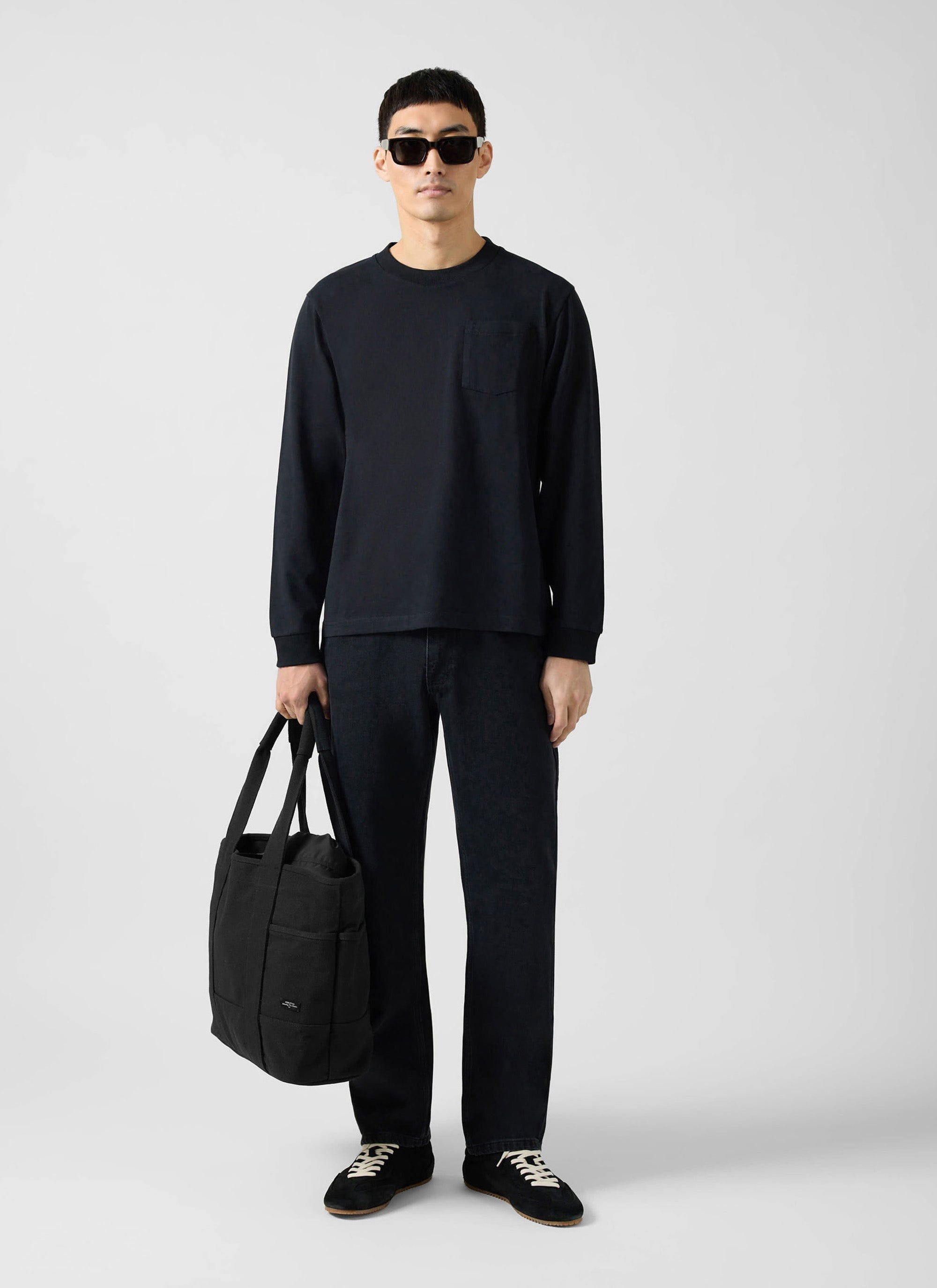 A model wears the Percival Long Sleeve Pocket T Shirt in black, styled with black jeans, trainers, and a tote bag.