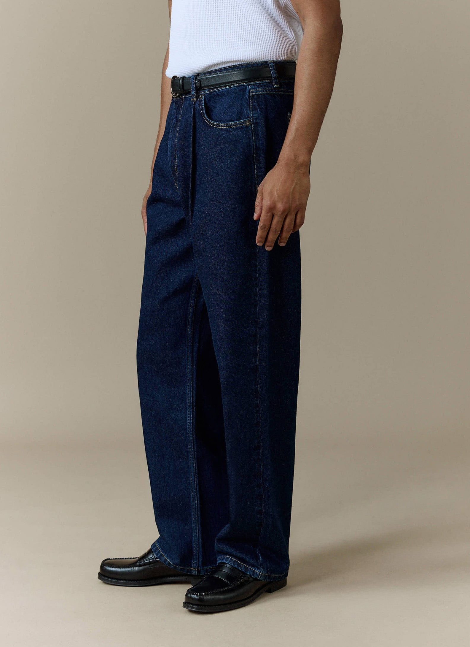A model wears the Percival Pleated Denim Jeans in dark indigo, styled with a white t-shirt, black belt, and black loafers.