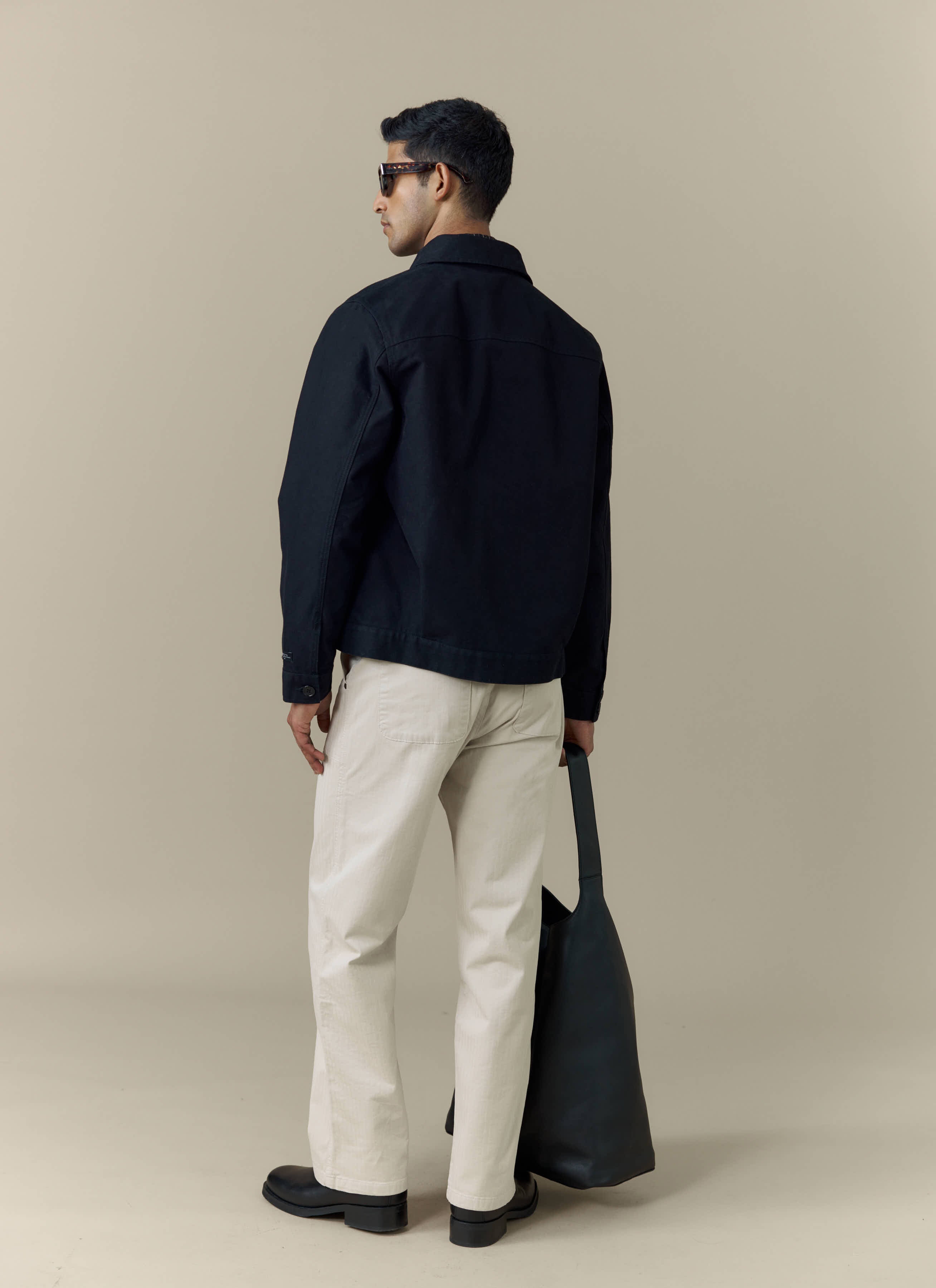 A model seen from behind wearing the black Ernst Jacket from Percival, styled with cream trousers and sunglasses.