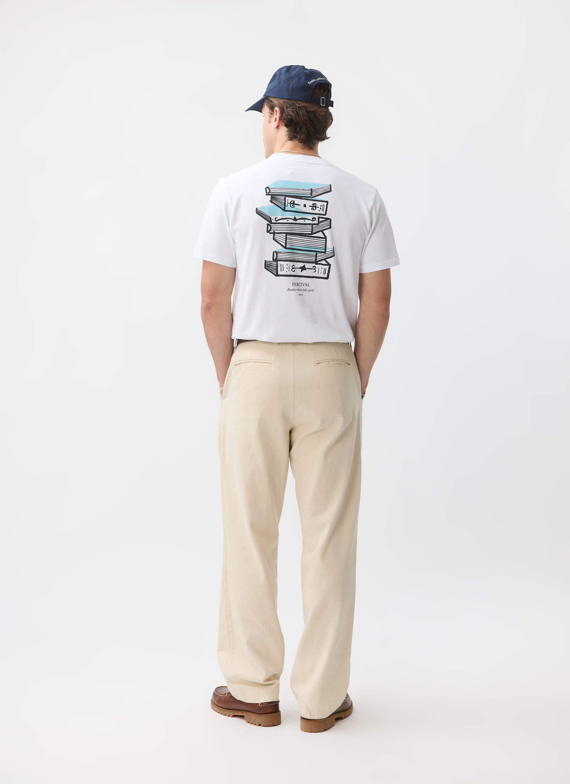 A model from behind wearing the Percival Duke T-shirt in white, showing the 'Routine Critical Daily Goods' book graphic.