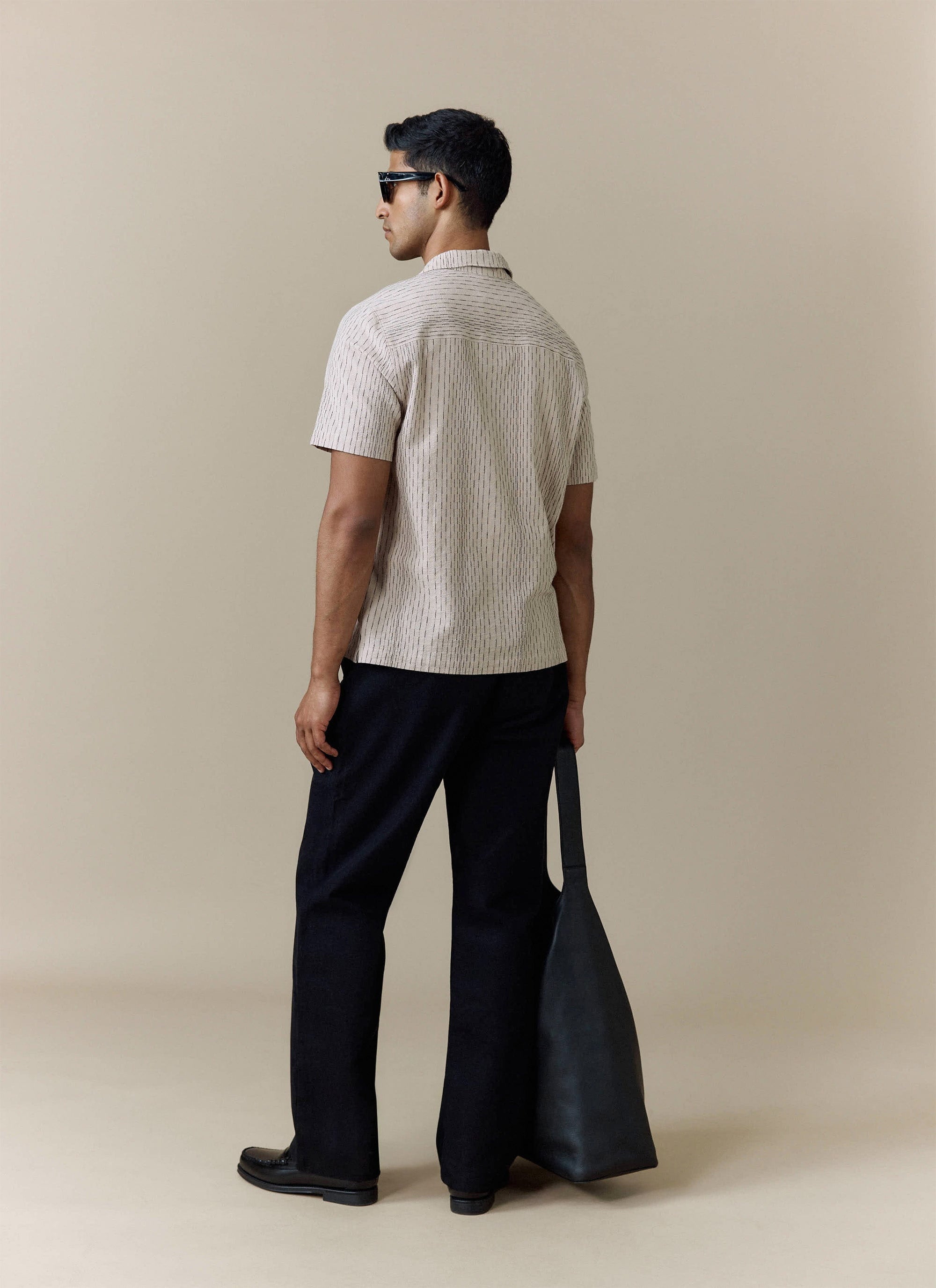 Rear view of the cream Percival Leland Cuban Shirt, styled with black trousers and a large leather tote bag.