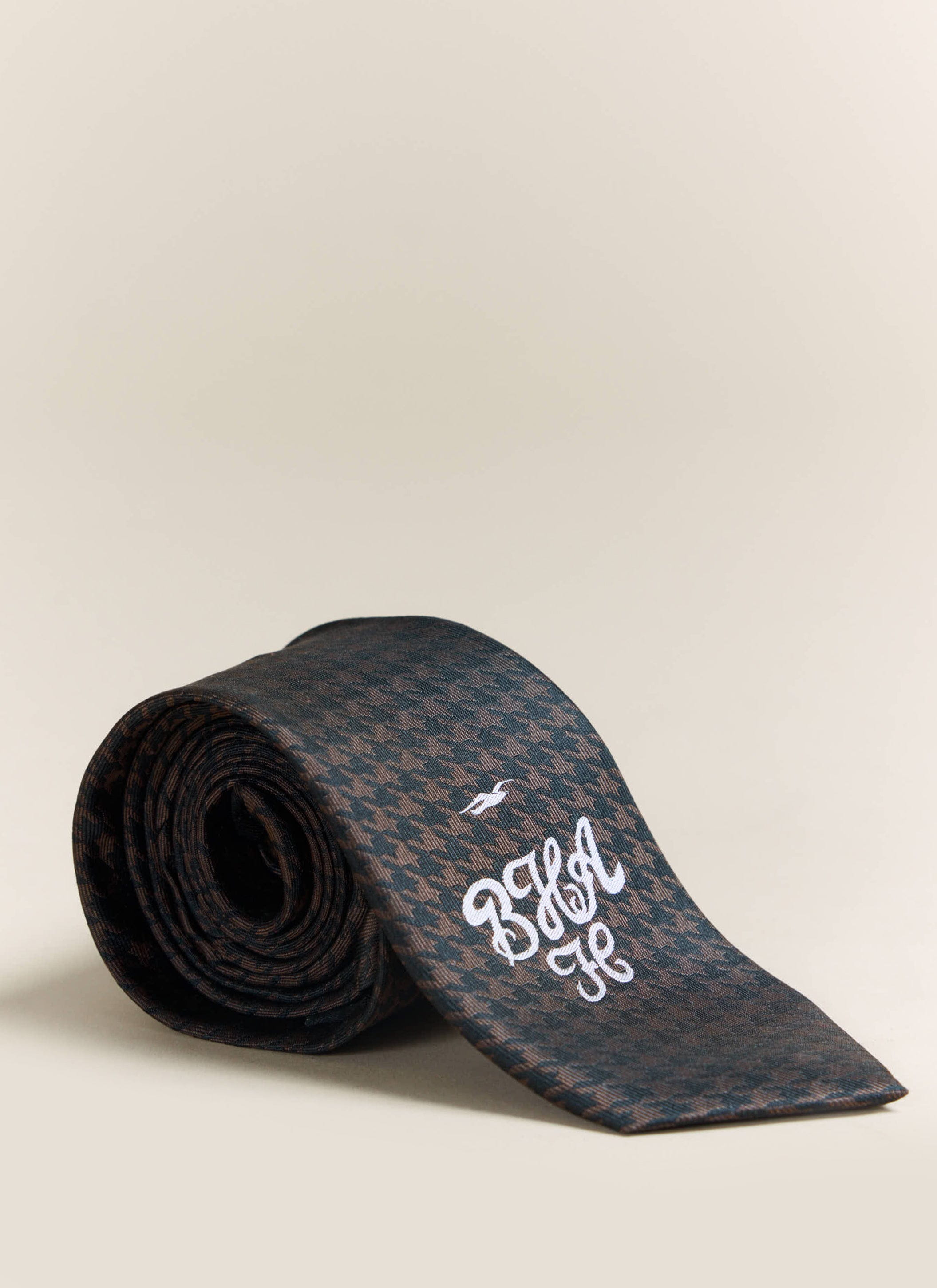 A rolled up Percival x Brighton & Hove Albion F.C. tie in brown silk, featuring a houndstooth pattern and embroidered logo.