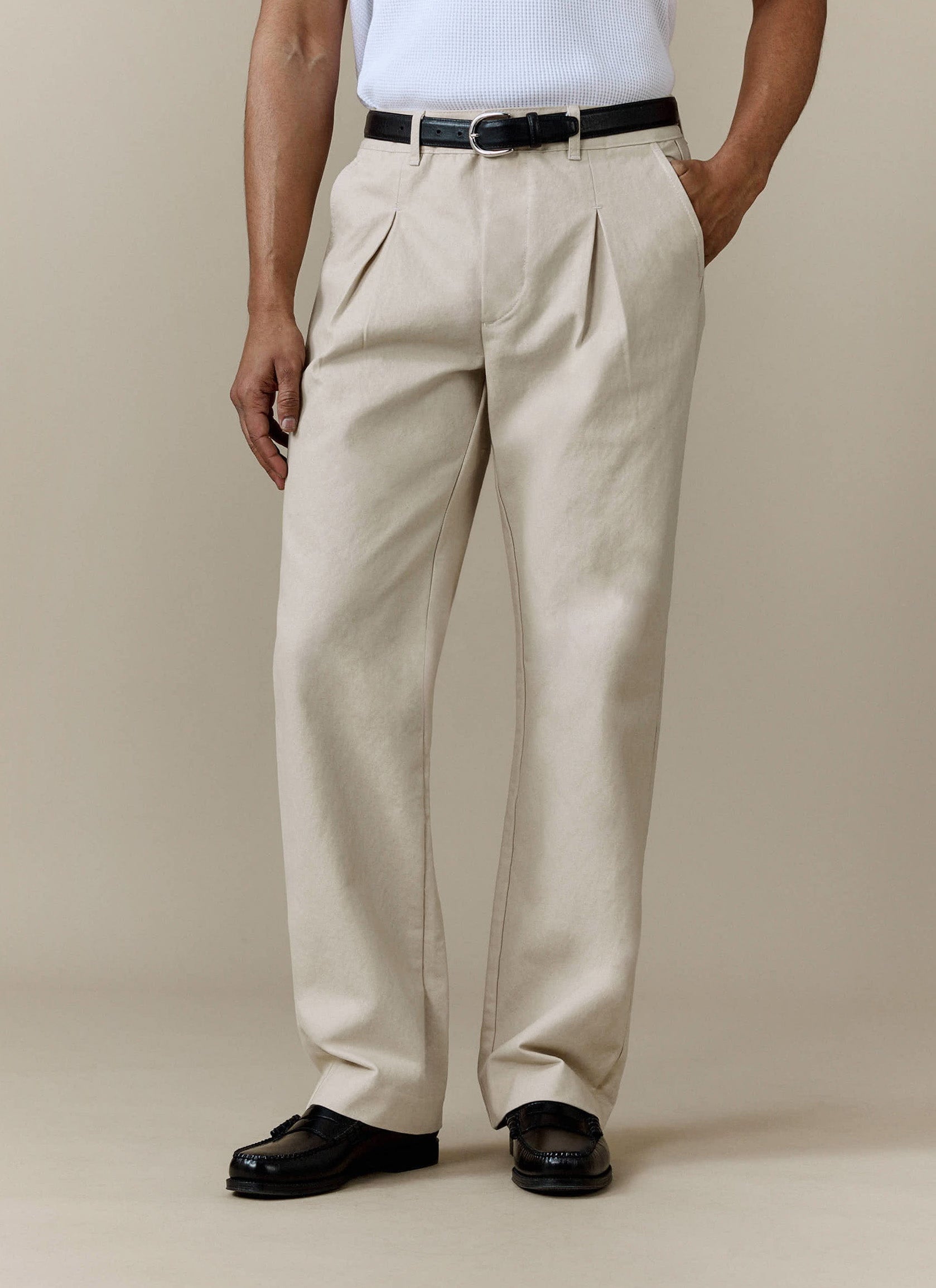 A model wears the ecru Seb Inverted Pleat Trousers by Percival, styled with a black belt and black loafers.