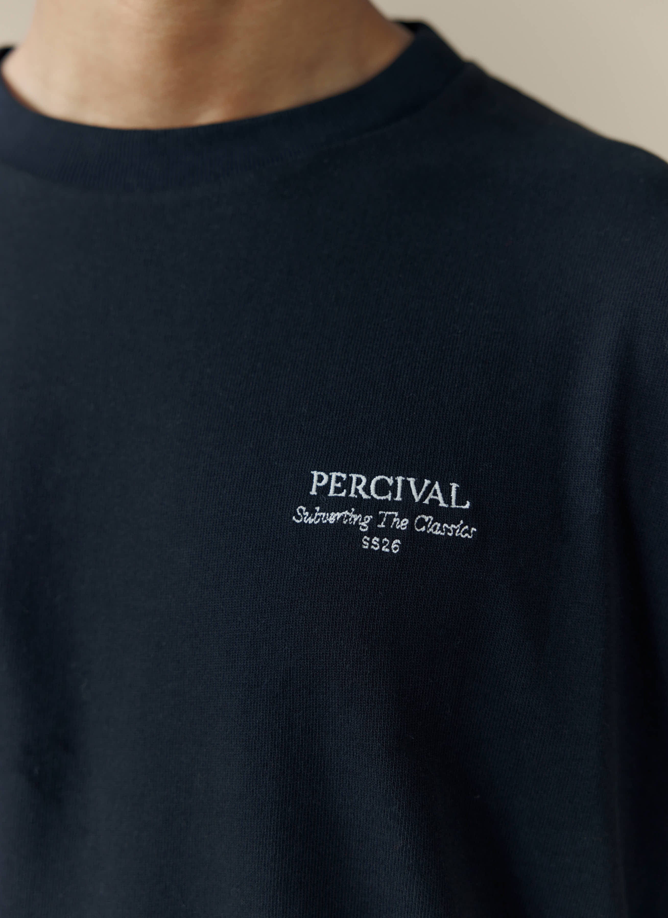A close-up of the white 'Subverting The Classics SS26' embroidery on the chest of a black t-shirt from Percival.