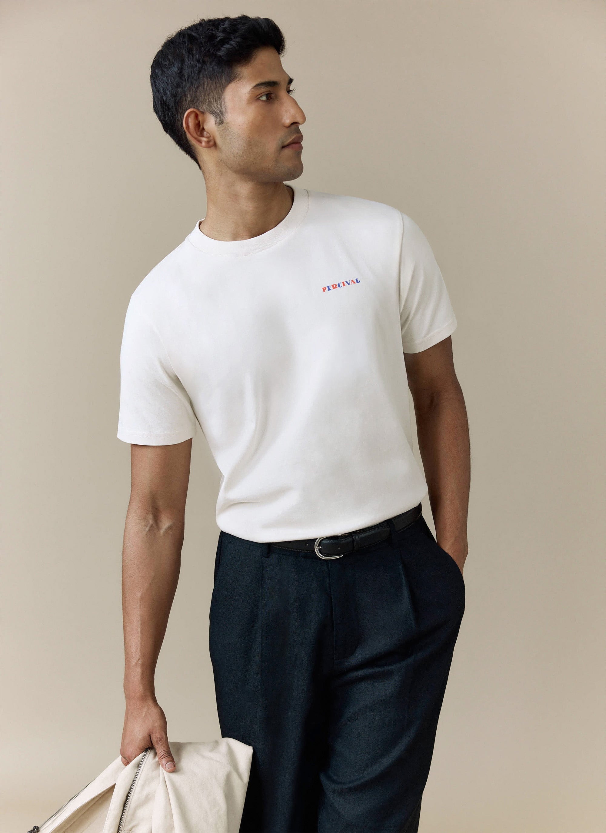 A man models the Percival logo t-shirt in ecru organic cotton, styled with dark pleated trousers.