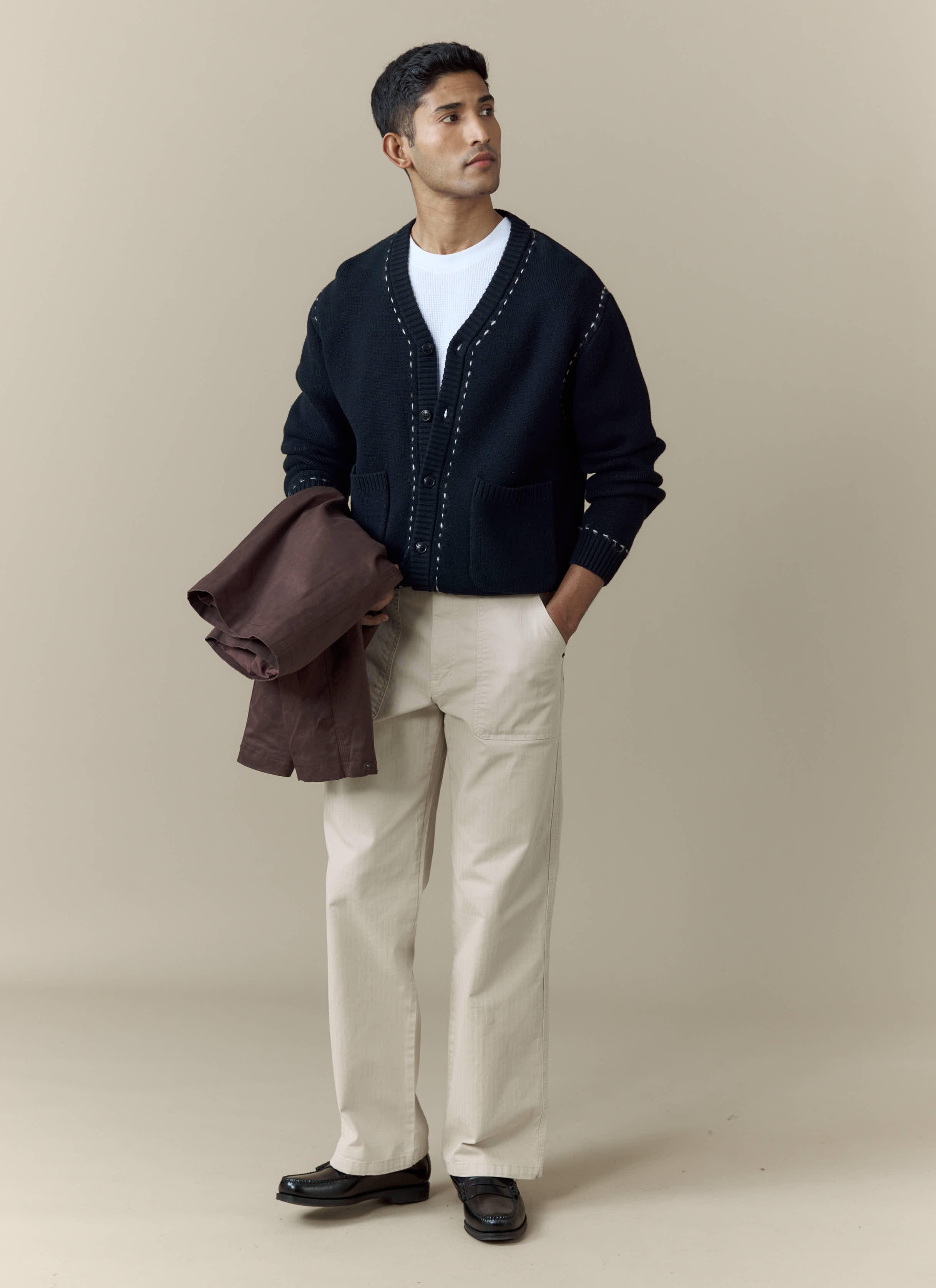 A model wears the black Module Boxy Cardigan from Percival, styled with beige trousers and a white t-shirt.