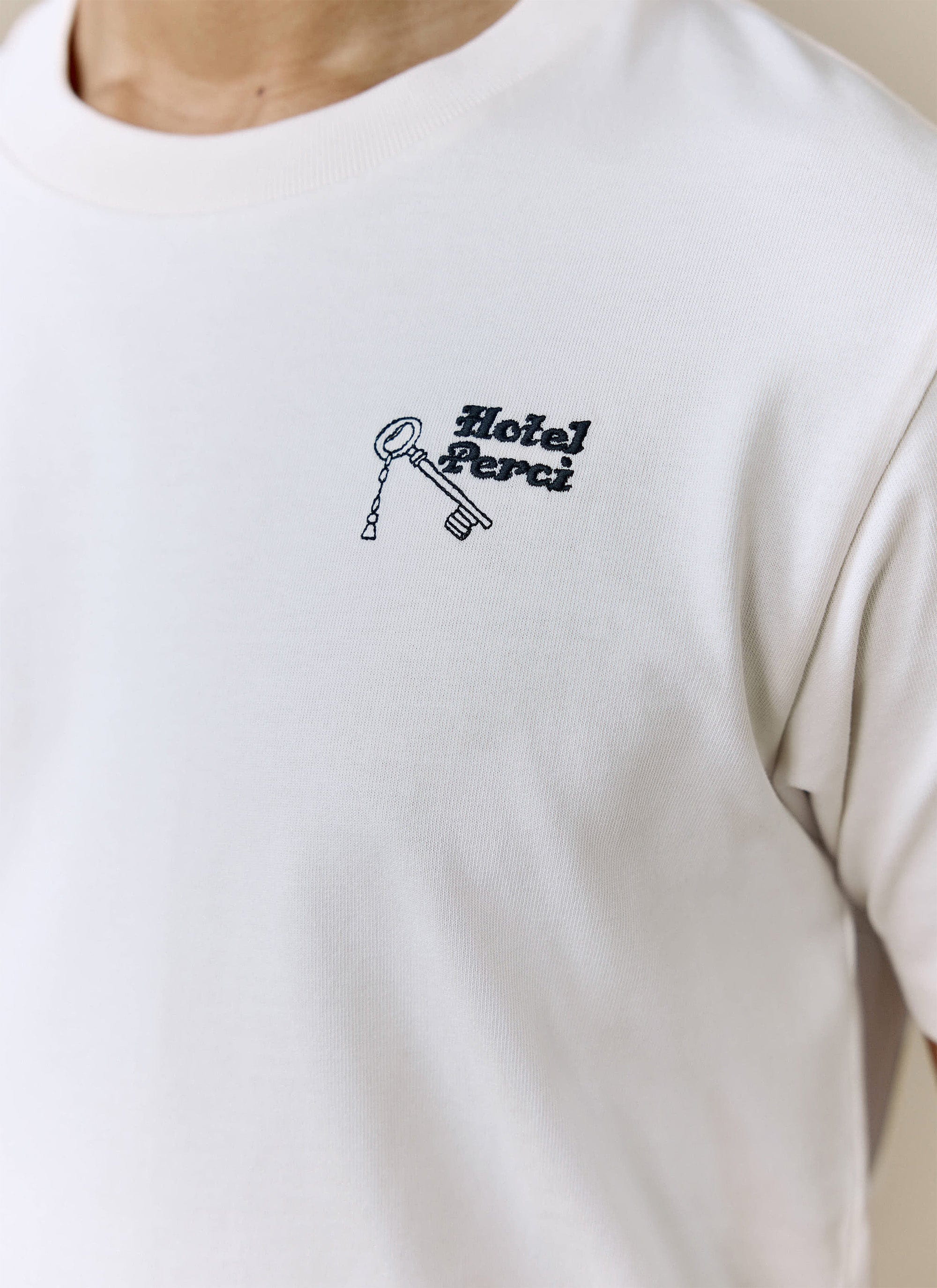 A close-up of the embroidered 'Hotel Perci' key graphic on the chest of the ecru Hotel T Shirt from Percival.