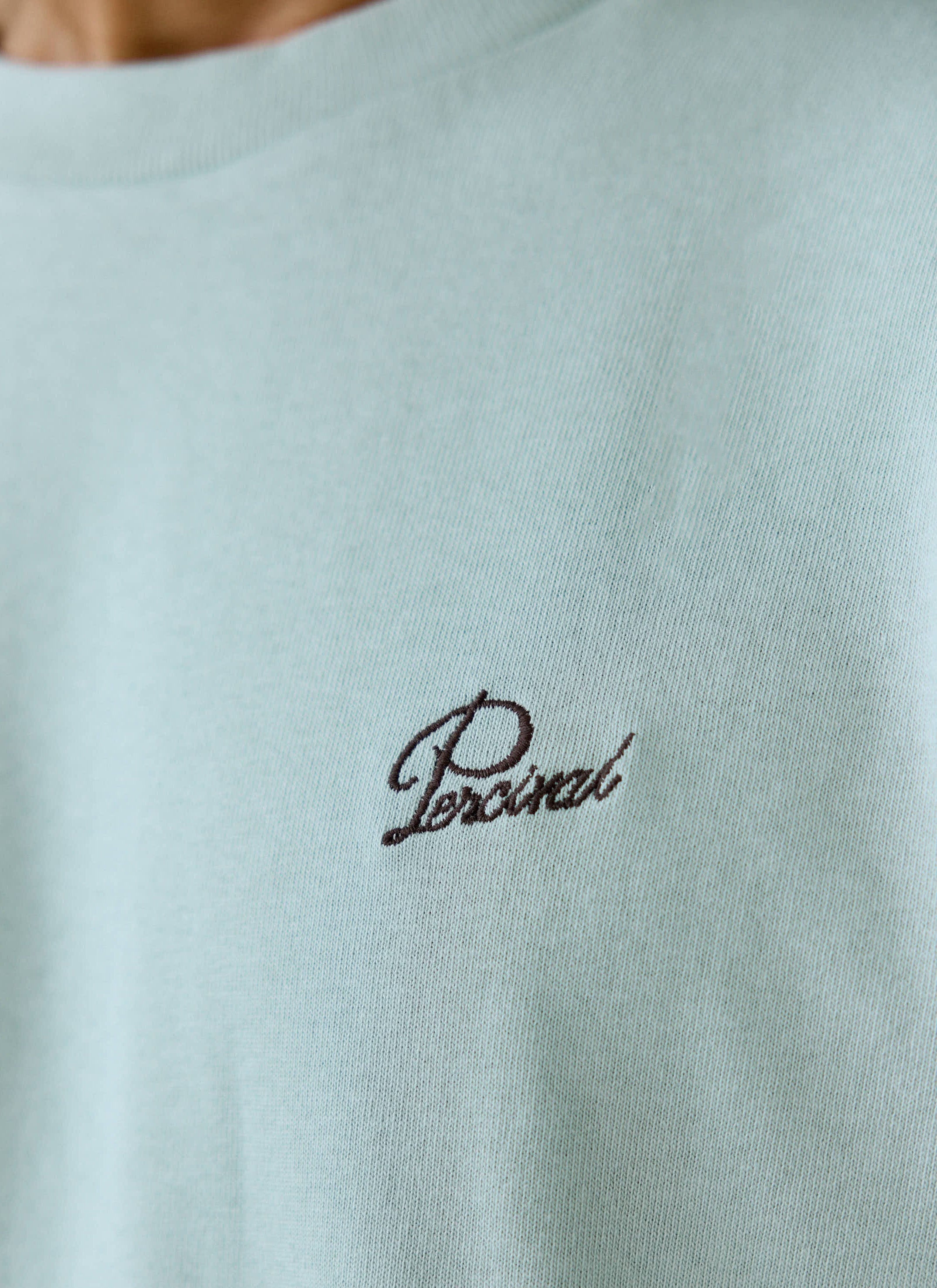 A close-up of the dark script embroidery on the chest of the sage green Perci Script T Shirt from Percival.