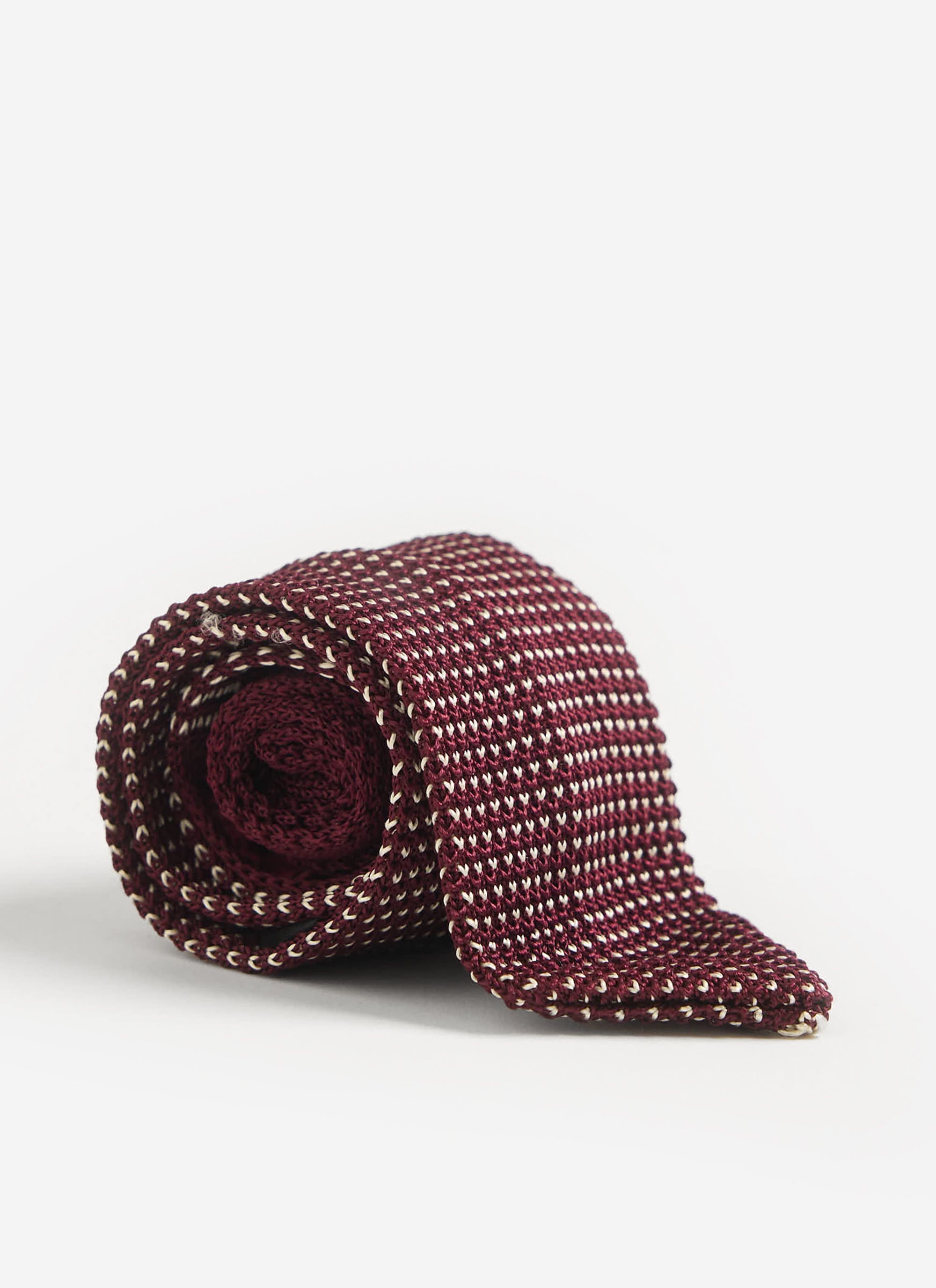 The Percival Birdseye Silk Tie in red, rolled neatly to showcase its intricate knit texture on a white studio background.