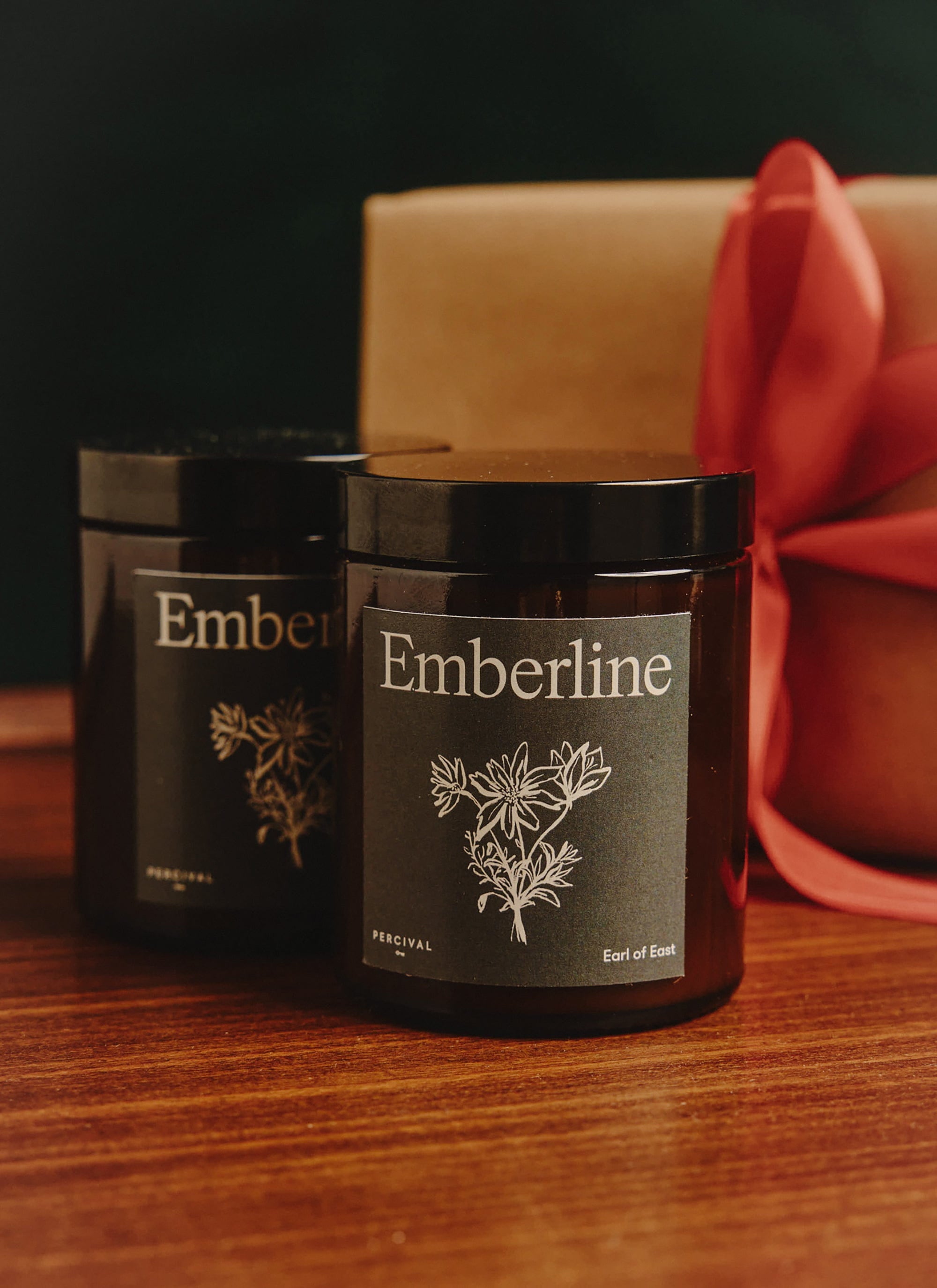 Two Percival x Earl of East Emberline candles in amber jars on a wooden table, with a gift box and red ribbon behind.