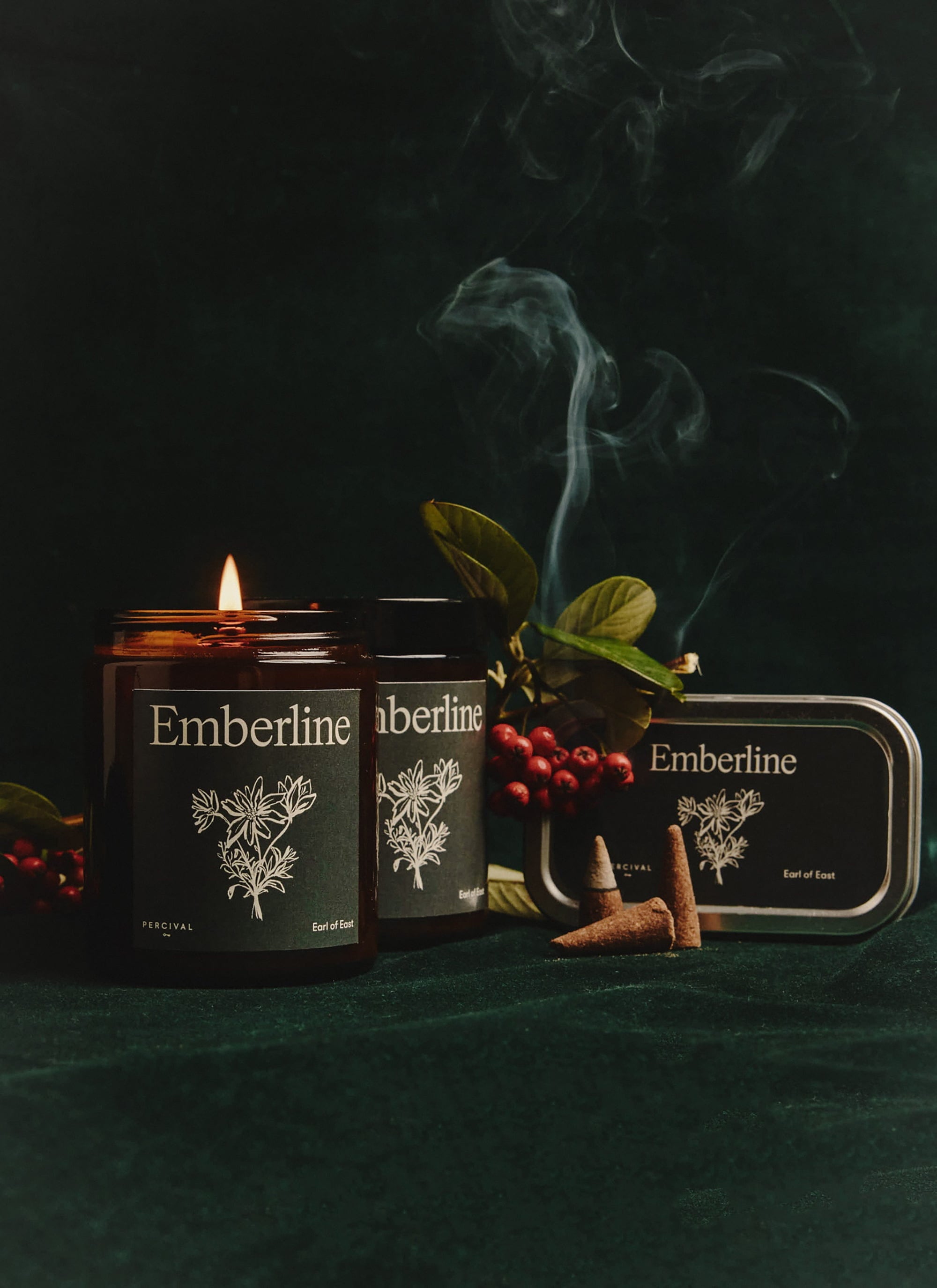 The Percival x Earl of East Emberline candle set, with a lit candle, incense cones and berries on a dark green background.
