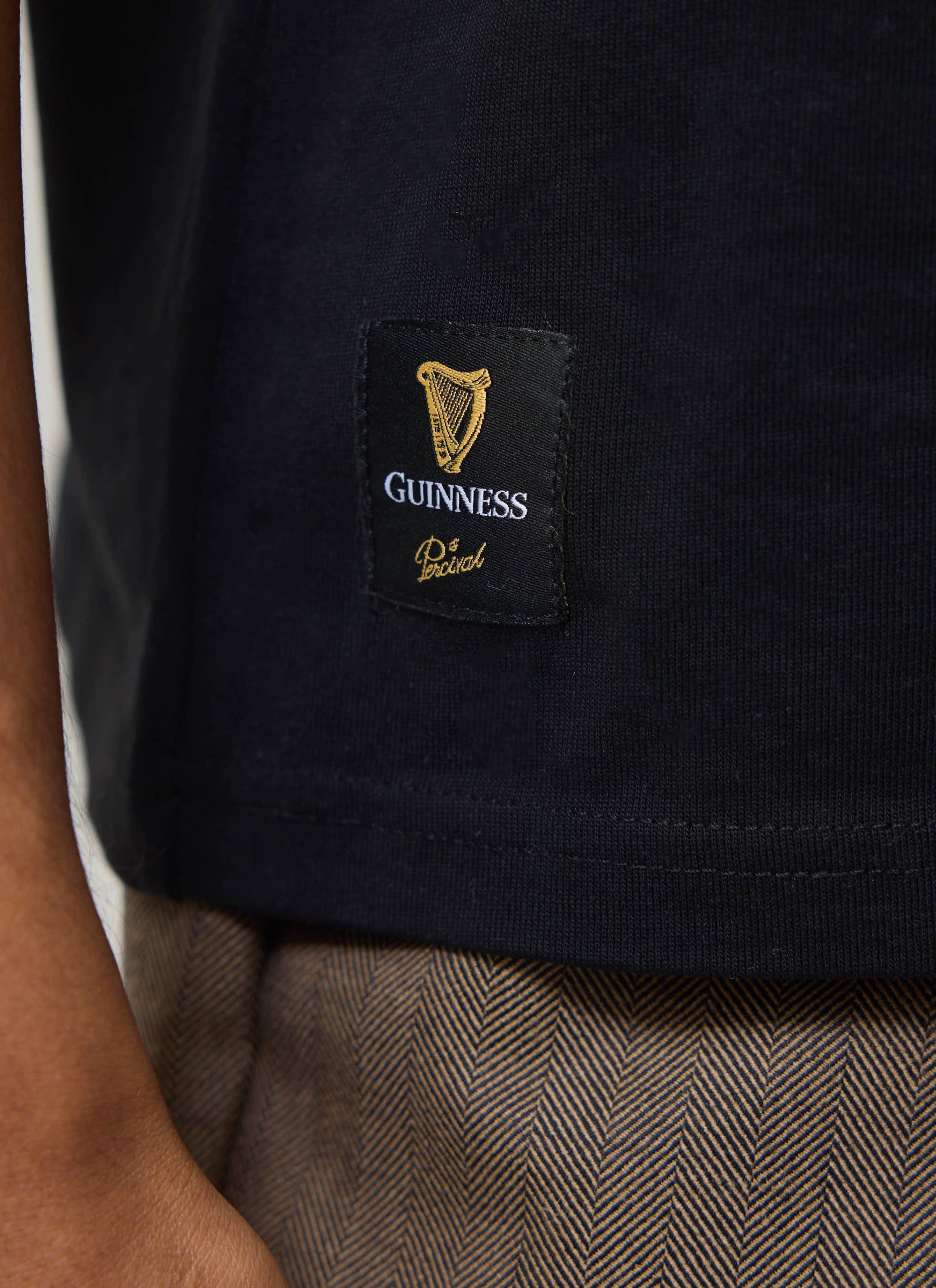 A close-up shot of the woven label on the hem of the black Percival x Guinness Stout T-shirt, showing the gold harp logo.