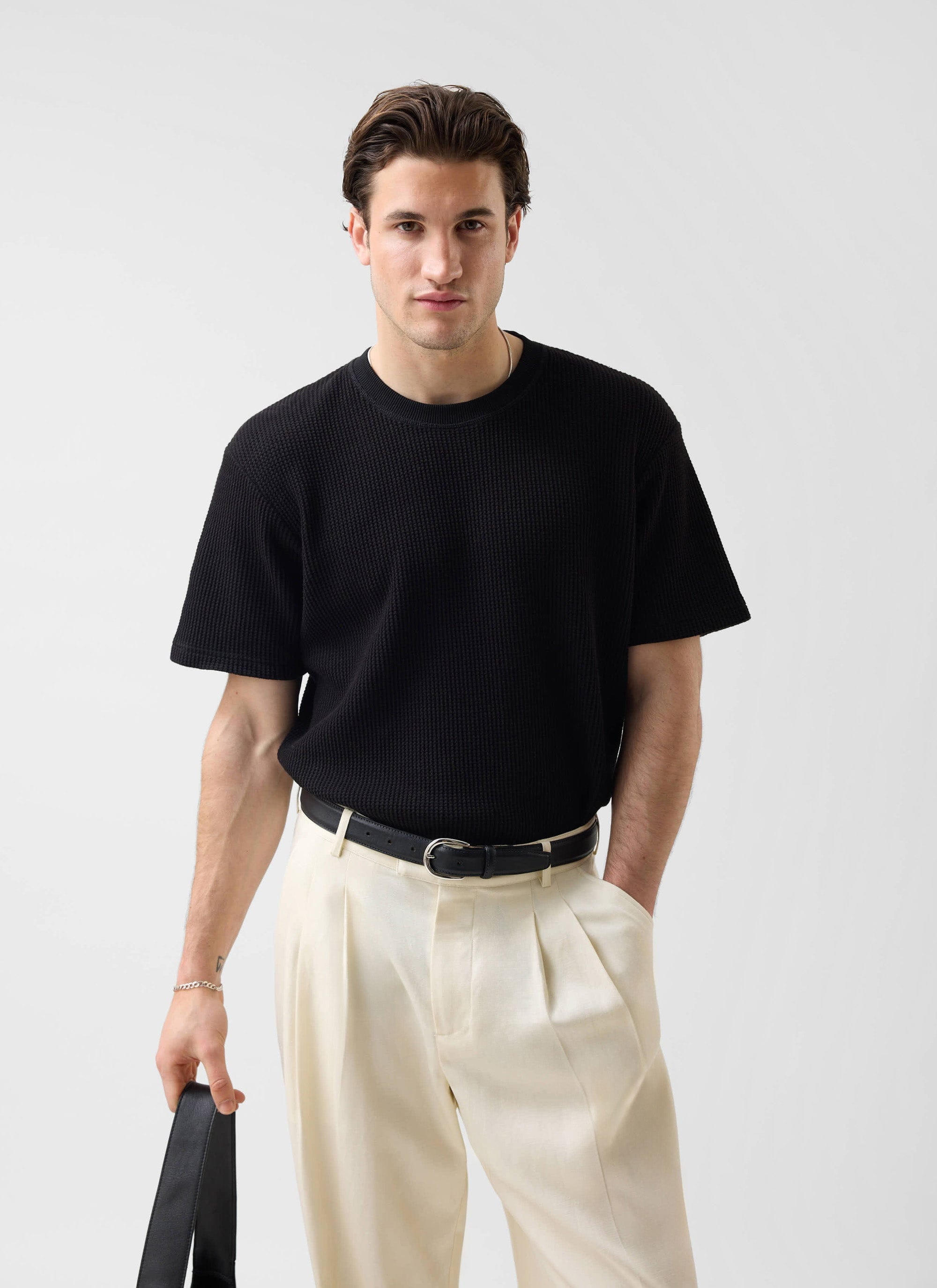 A model wearing the Percival Waffle Short Sleeve T-Shirt in black, paired with smart cream pleated trousers.