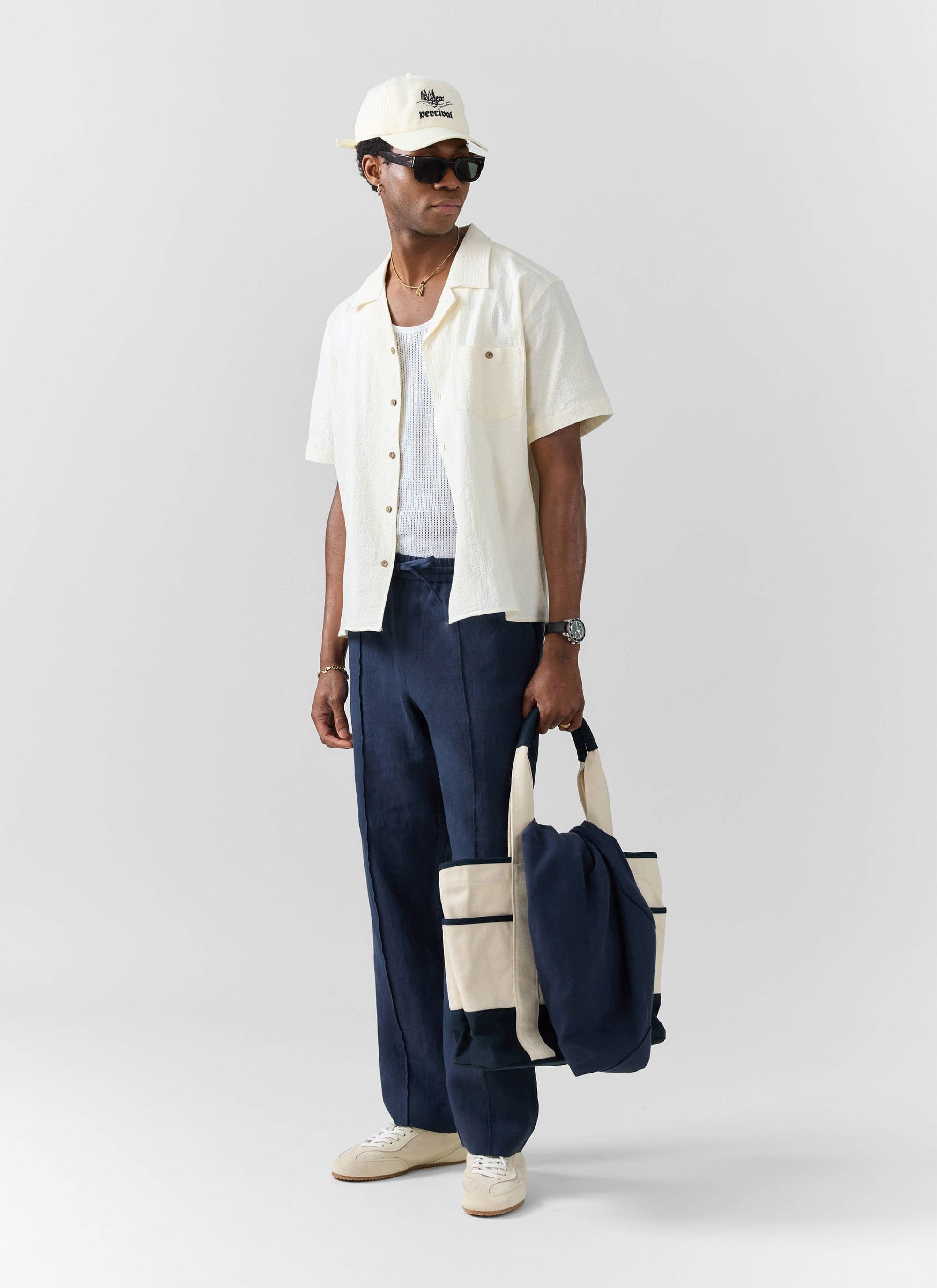 A full-length shot of a model wearing the Percival Lorena Wide Leg Trousers in navy linen, styled with a cream shirt and cap.