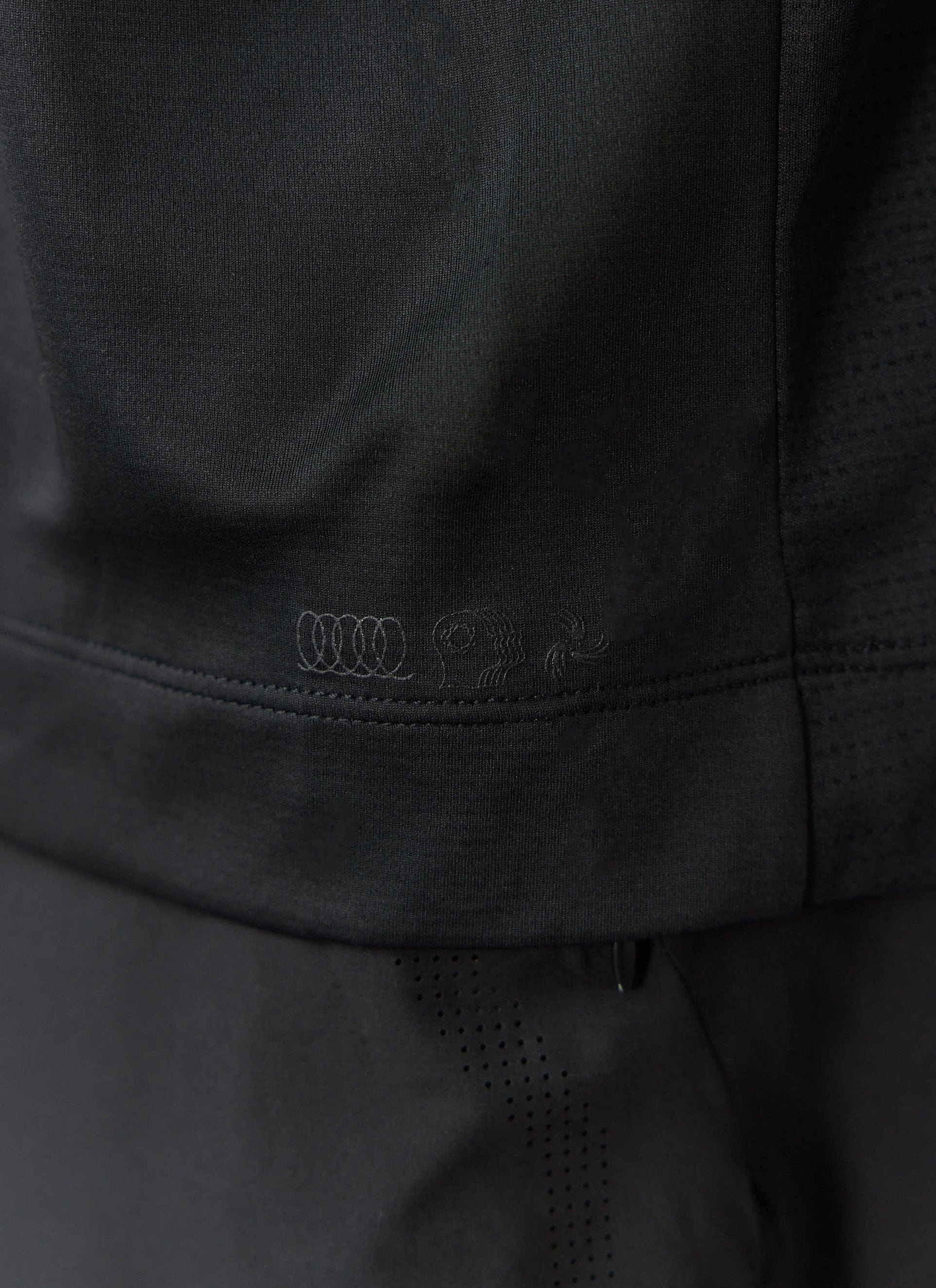 A close-up of the Percival PCVL Performance Top in black, showing the subtle tonal graphic print on the hem.