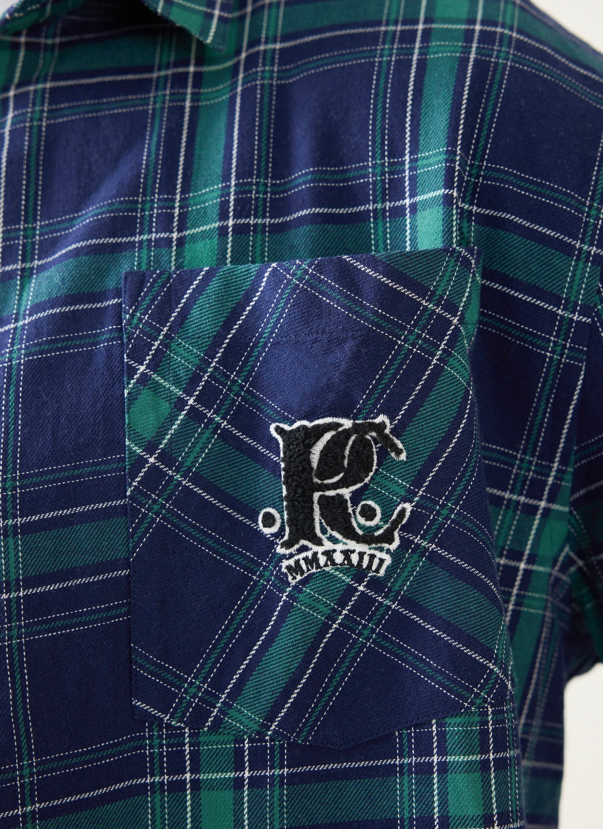 A close-up of the chenille 'P' logo on the chest pocket of the Champion x Percival blue and green tartan shirt.