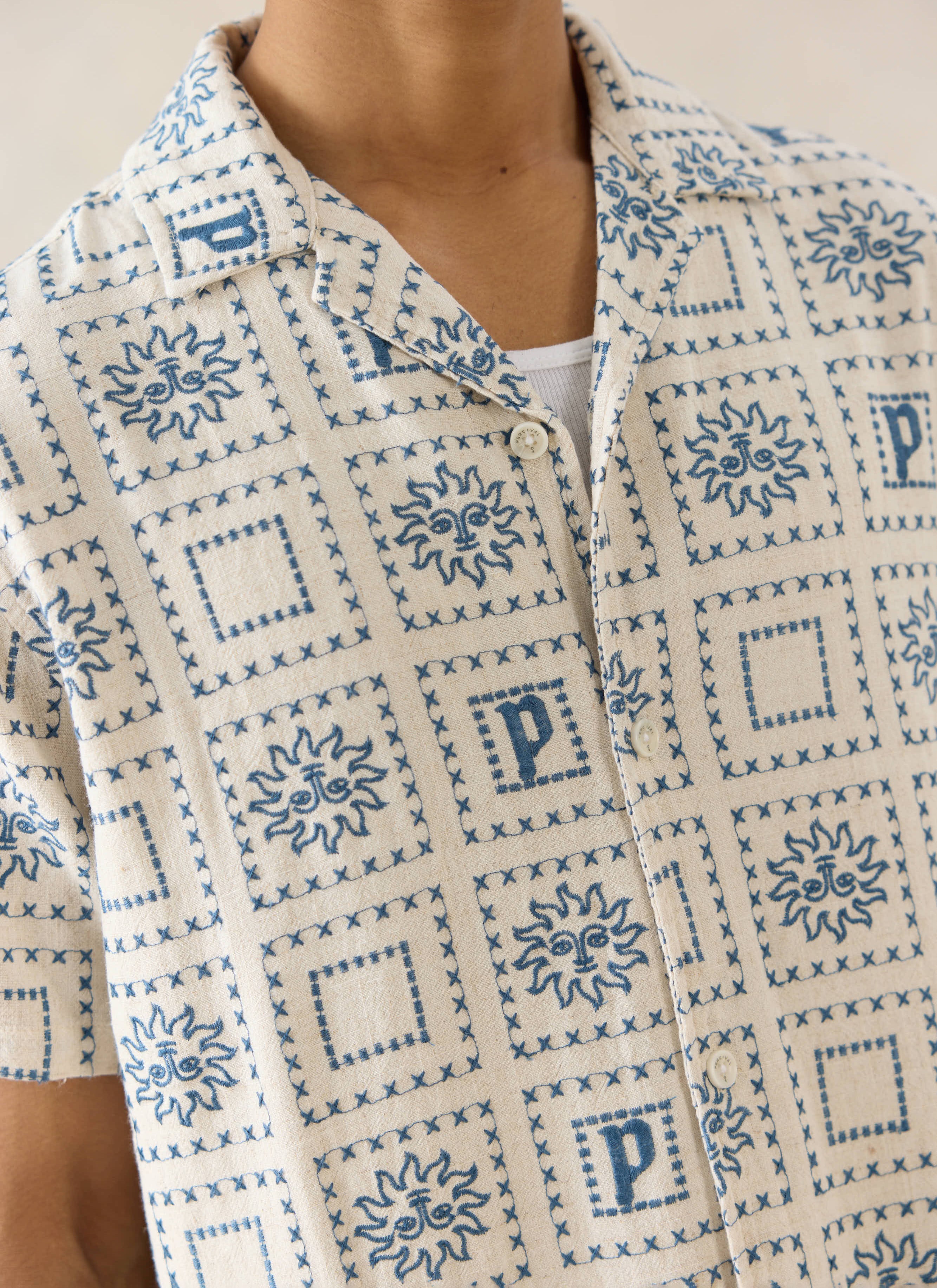 The Percival Solis Heaton Cuban Shirt in cream, a linen-blend top with blue sun motifs and 'P' embroidery on a model.