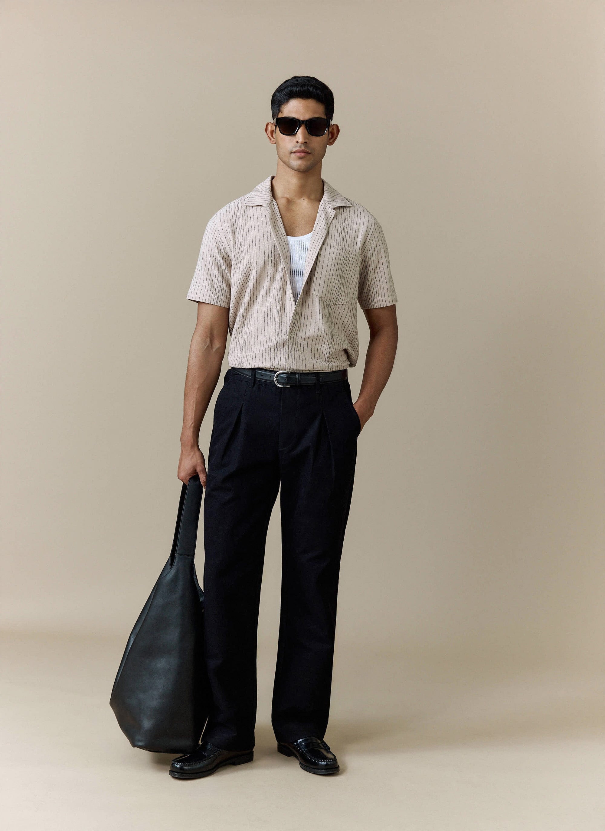 A model wears a Percival pinstriped short-sleeve shirt, black trousers, and sunglasses, holding a black tote bag.