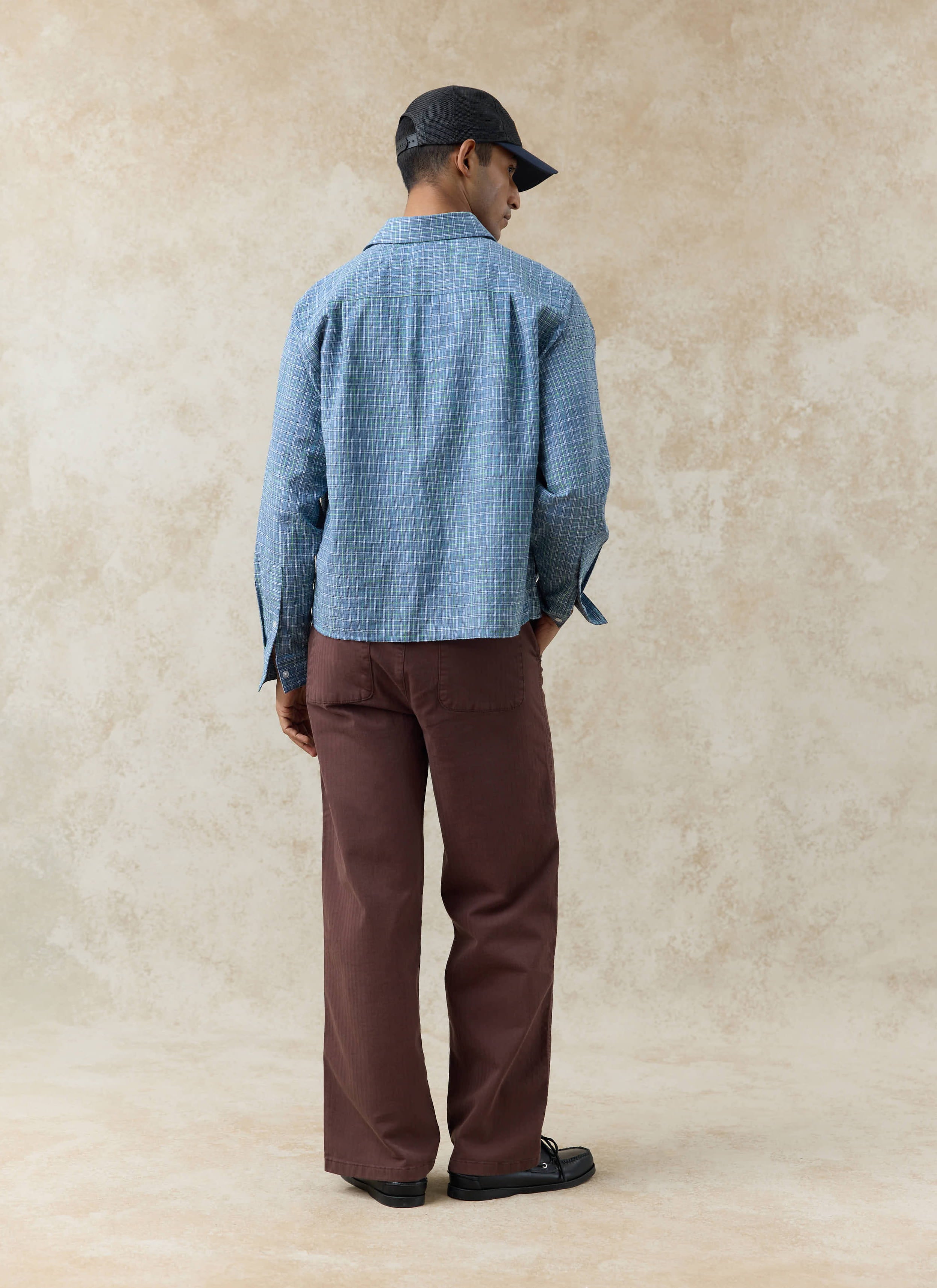 Rear view of the blue cotton Percival Hinsley Checked Boxy Shirt, styled with brown trousers and a black cap.