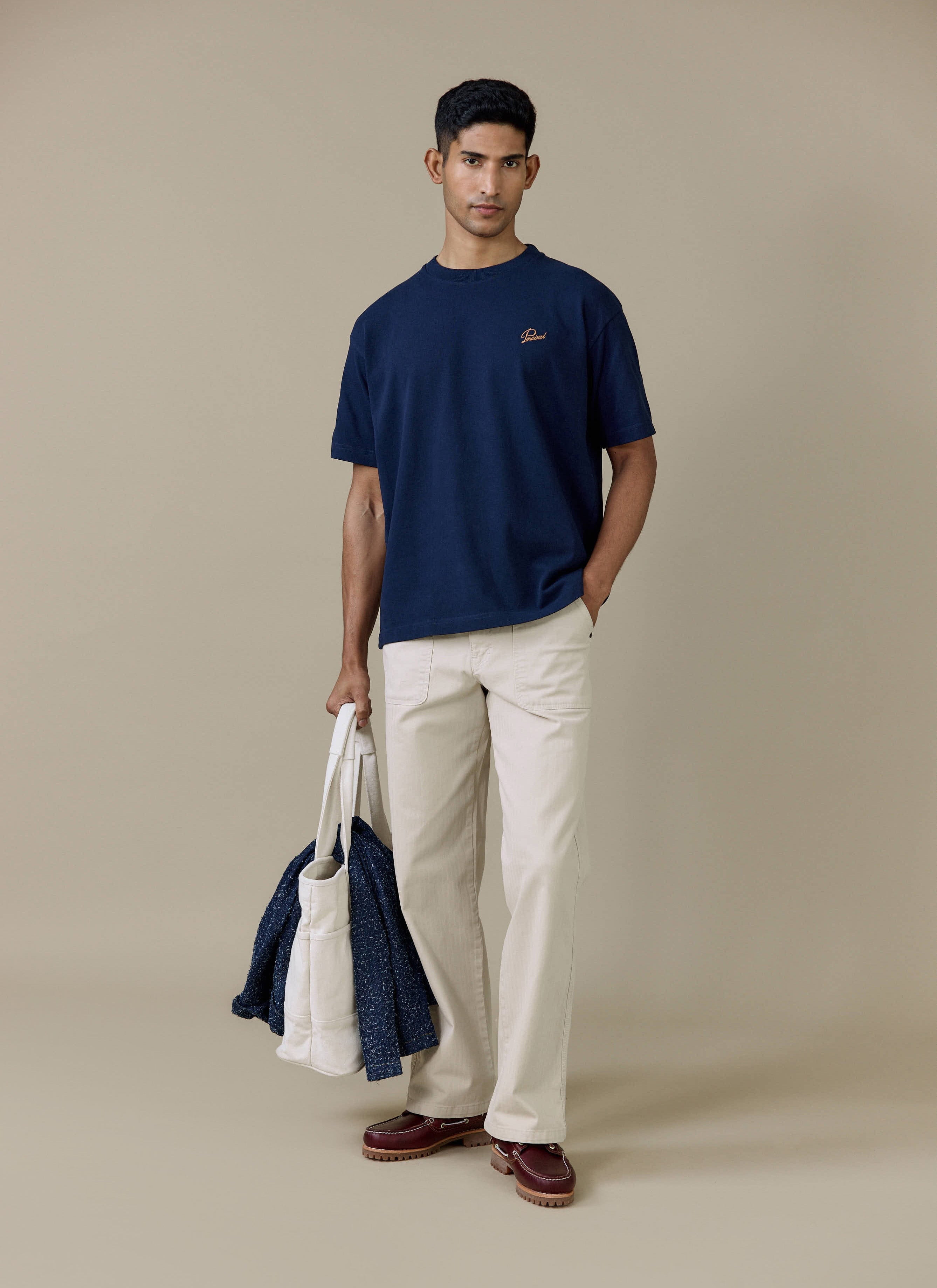 Model wearing the navy Percival Perci Script T Shirt in organic cotton, styled with cream trousers and a white tote bag.