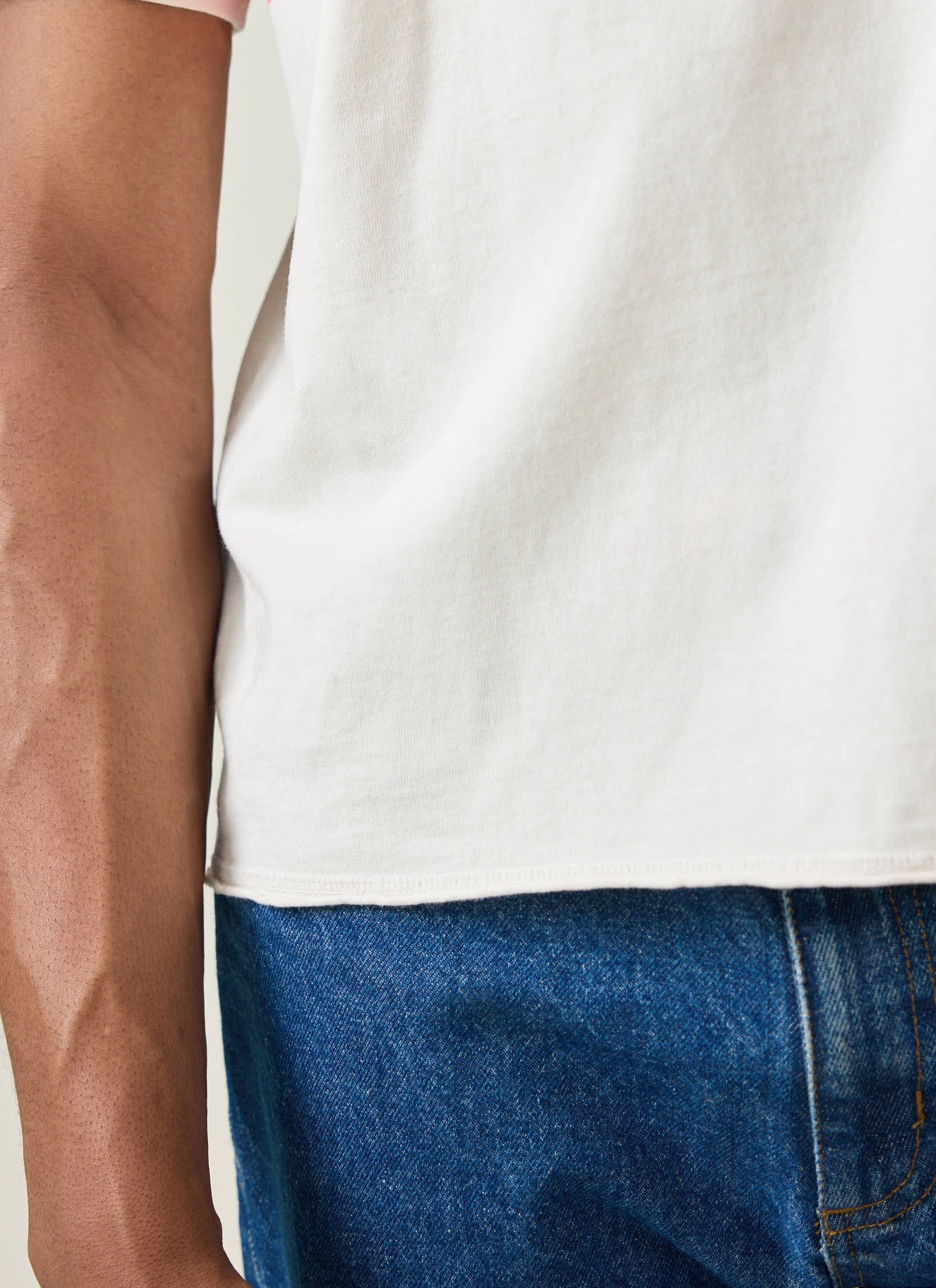A close-up of the raw, undone hem of the Champion x Percival Flag Stripe T-shirt in ecru, worn with blue jeans.