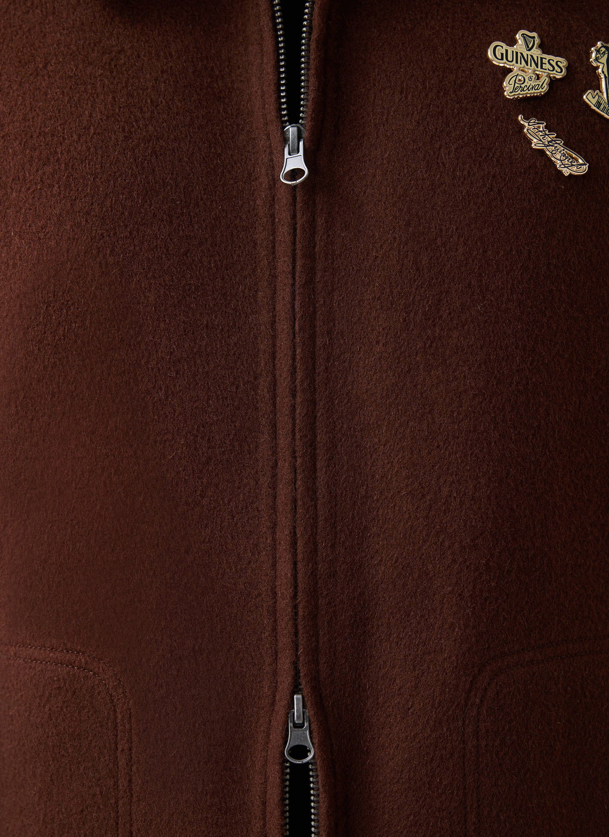 A close-up of the Guinness x Percival Wool Jacket in espresso brown, showing the zip and collaboration pin badges.