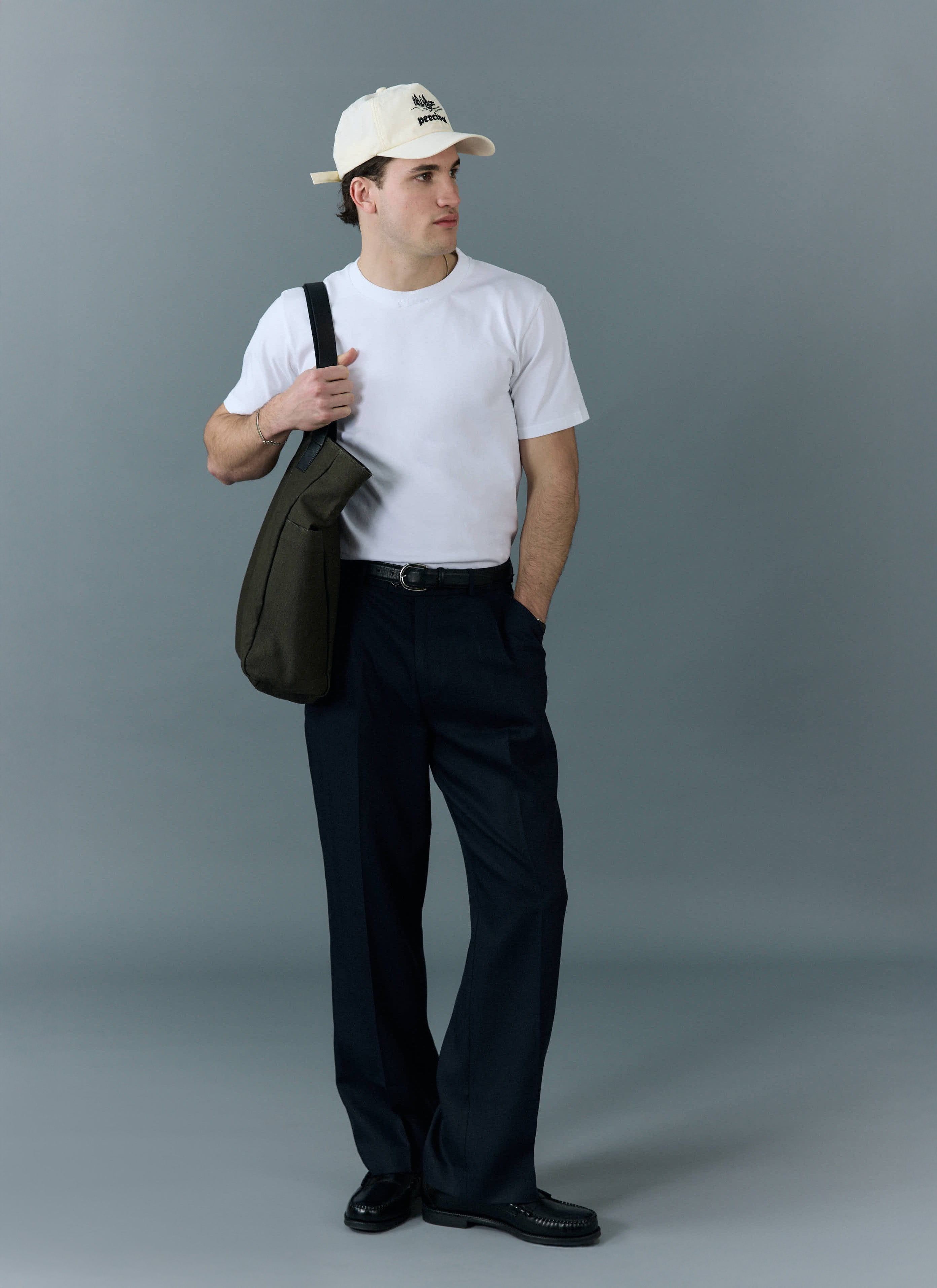 A man wearing a Percival plain white t-shirt, dark trousers, a cap, and carrying a tote bag against a grey background.