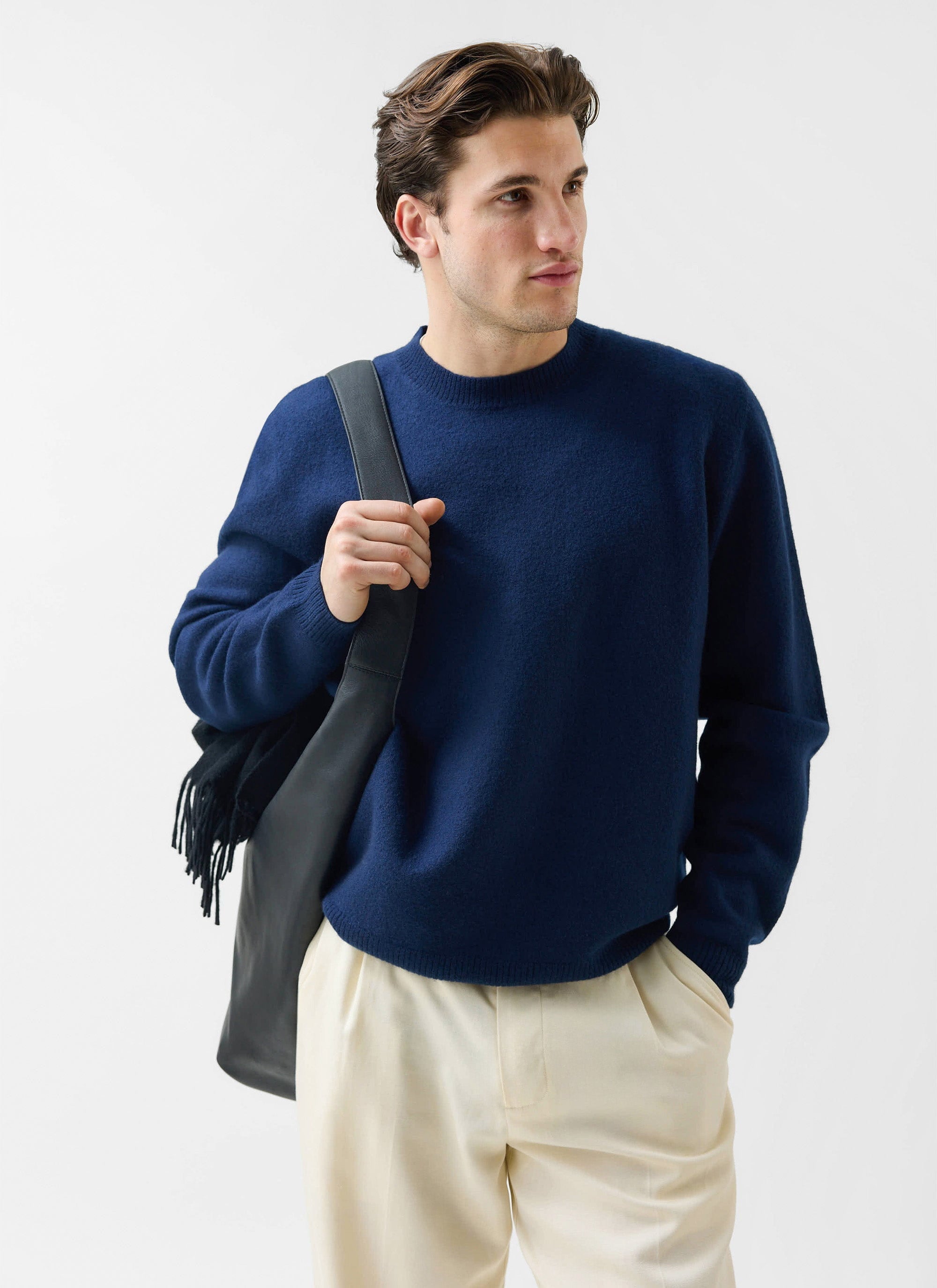 A man models the Percival Felted Regular Jumper in navy merino wool, styled with cream trousers and a black shoulder bag.